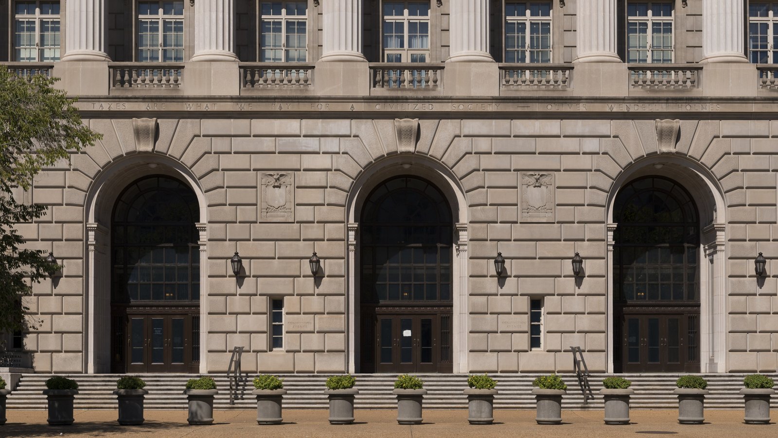 Three tall arched entrances in the pale limestone front of the Internal Revenue Service Building in Washington, photographed square on in daylight. A carved inscription runs in a band above them reading that taxes are what we pay for a civilized society, attributed to Oliver Wendell Holmes. Fluted pilasters and a stone balustrade sit above, small planted urns stand in a line along the pavement below, and nobody is in the frame.