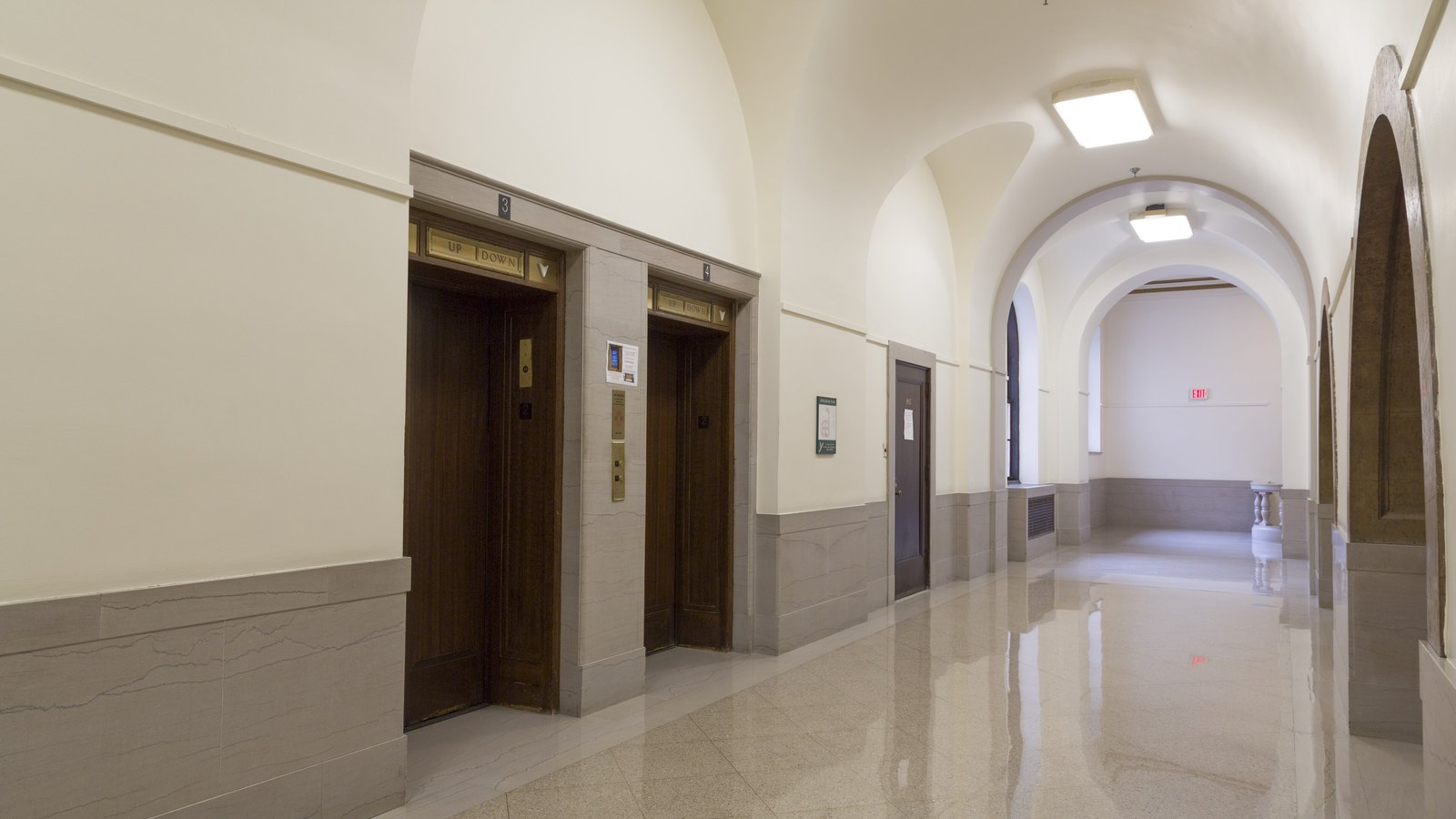 A vaulted, empty corridor inside the Internal Revenue Service Building in Washington, with two dark wooden lift doors numbered three and four, brass up and down indicators above them, marble wainscoting and a polished stone floor.