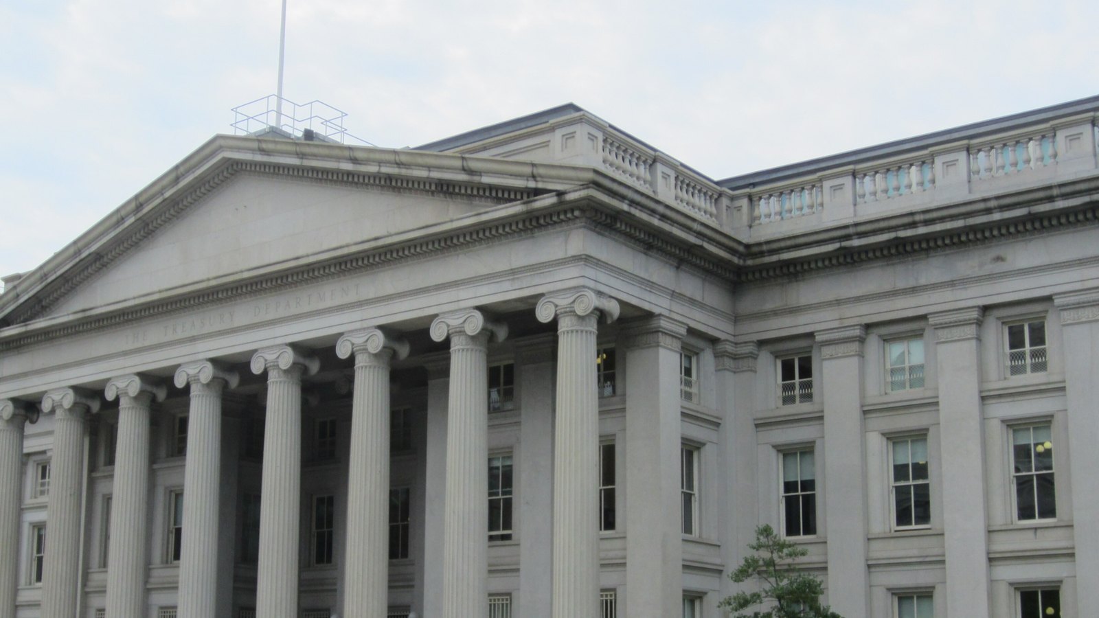 The pediment and Ionic columns of the Treasury Building in Washington, with The Treasury Department carved in the frieze, a rooftop balustrade and a pale overcast sky.
