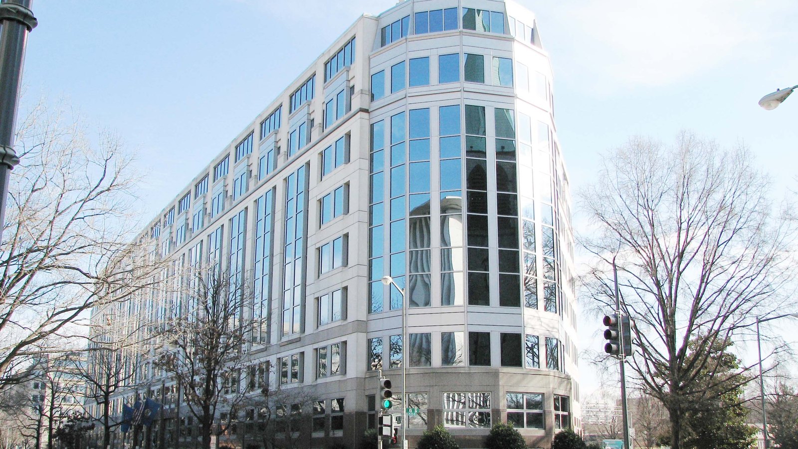 The United States International Trade Commission building in Washington seen from a street corner, a pale stone and blue glass office block rising to a rounded glazed turret, with bare winter trees, a traffic signal and a row of flags in front of it.