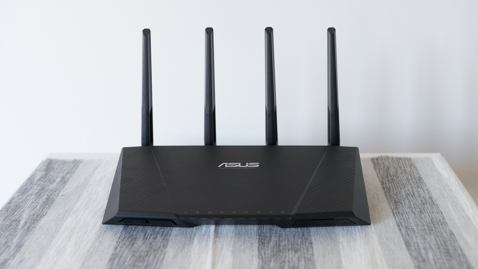 A black Wi-Fi router with four upright black antennas standing on a pale striped table against a plain white wall, photographed straight on. The manufacturer name ASUS is embossed on the front of the case and no other brand mark, person or place is in the frame.
