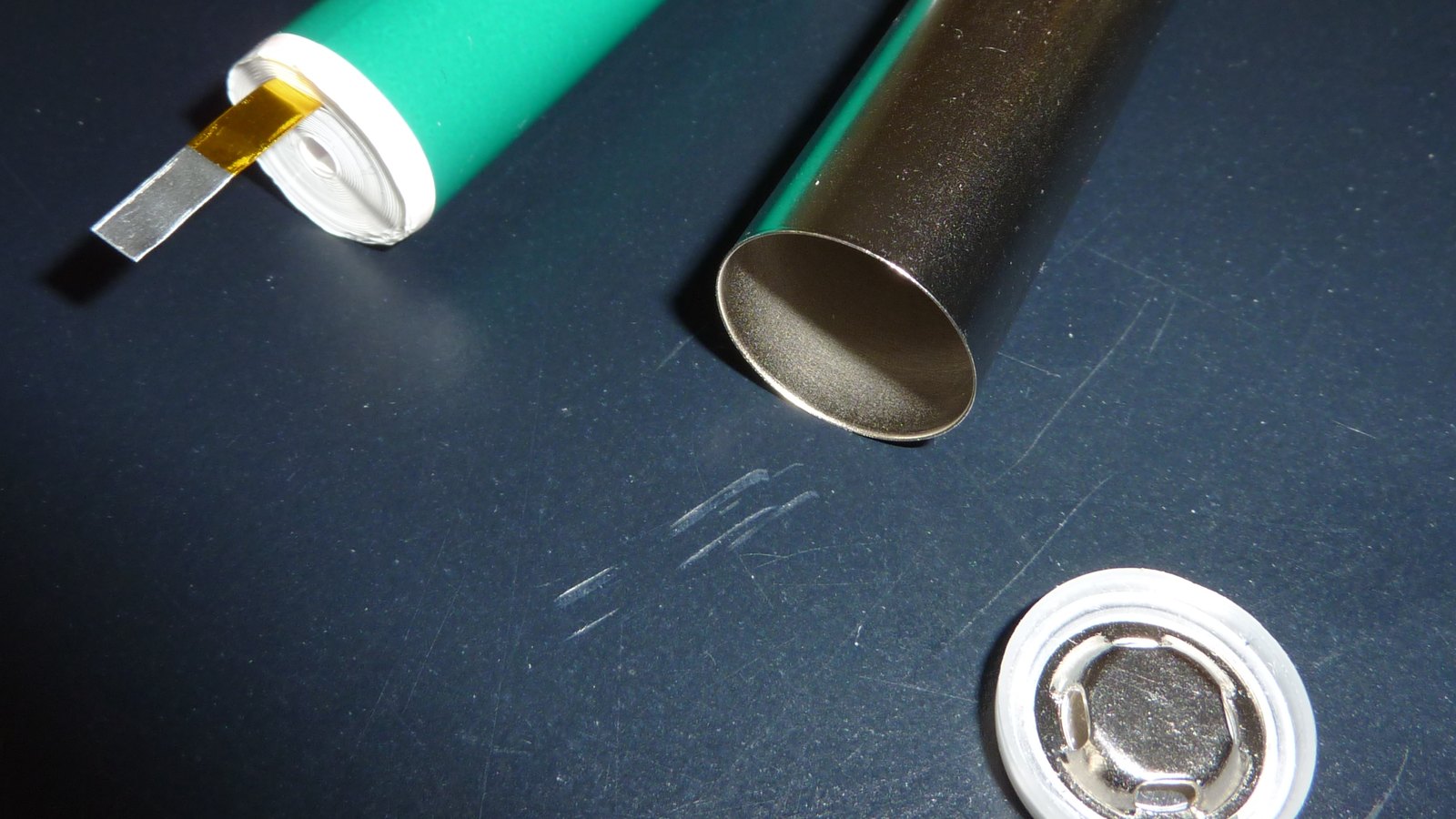 A cylindrical rechargeable battery cell taken apart on a dark bench: the tightly wound spiral electrode roll in a green wrapper with a bright metal tab folded out of one end, the empty polished steel housing lying open beside it, and part of the round metal top cap assembly at the lower edge. No text, brand mark, person or place is in the frame.