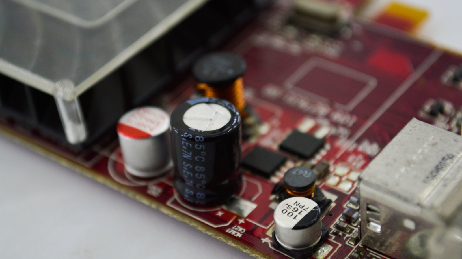 A close view of a dark red circuit board, with two electrolytic capacitors standing among small black surface-mount chips, an aluminium heatsink at the left edge and a metal USB socket at the right.