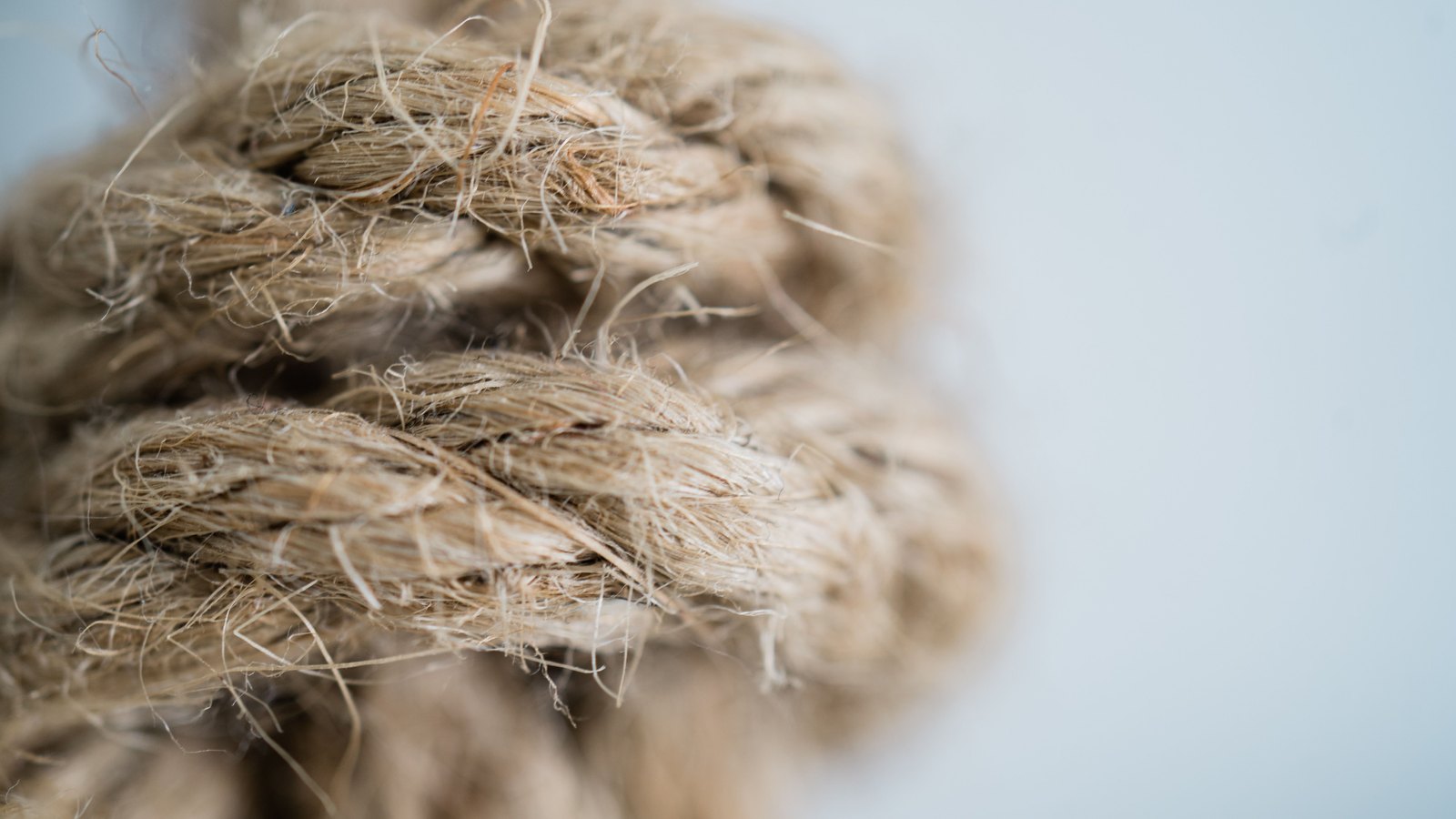 A close photograph of a coil of pale natural fibre rope, its three strands twisted in a tight spiral across the left two thirds of the frame, with loose hairs standing out from the surface. The background is plain and pale grey. No text, lettering, brand mark, person or place is in view.