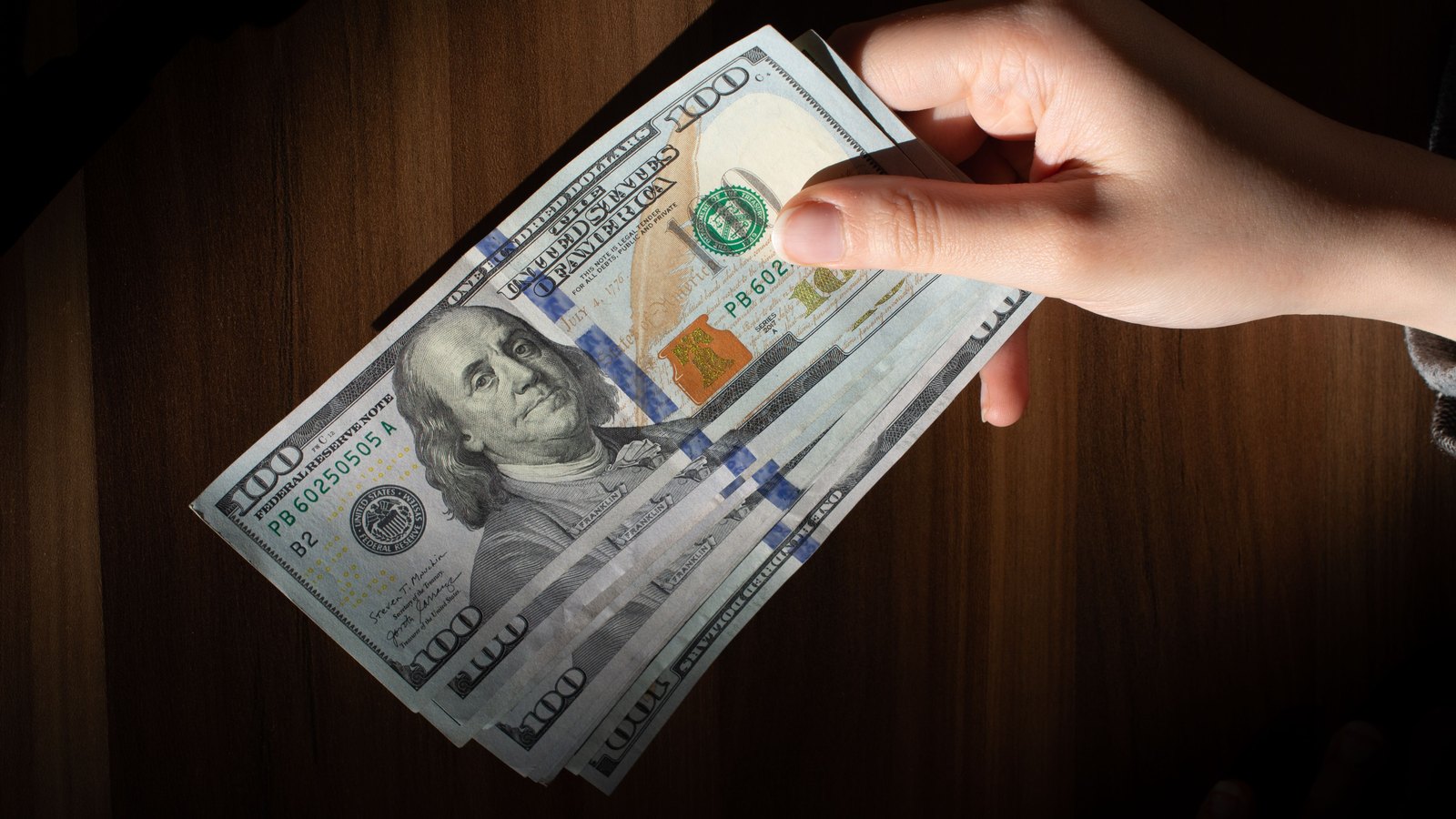 A hand holds a fanned stack of United States $100 banknotes above a wooden tabletop.