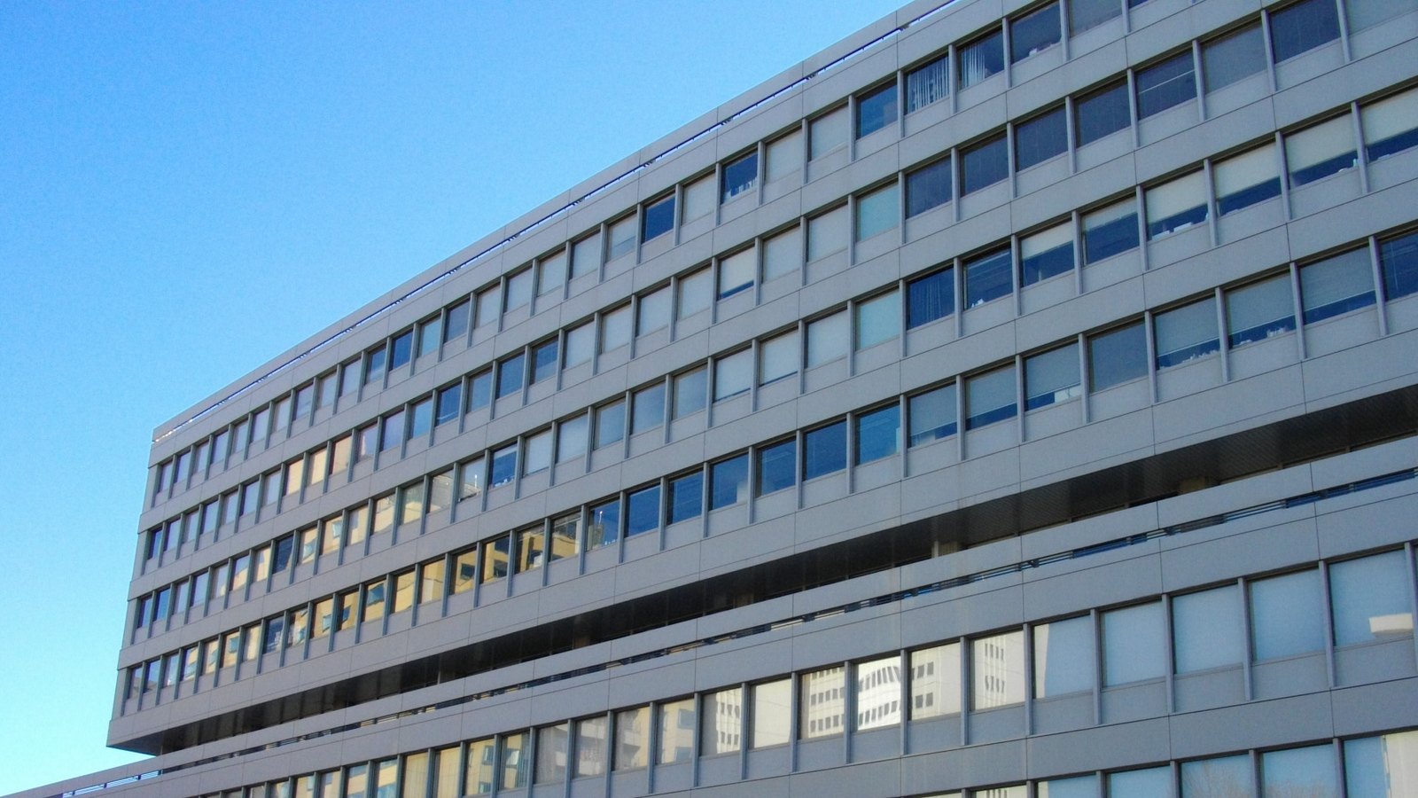 The curved grey facade of the Statistics Bureau of Japan building, its rows of windows running across the frame in stepped horizontal bands, neighbouring office blocks reflected in the glass, and clear blue sky at the upper left. No people, vehicles, signage or lettering are in view.