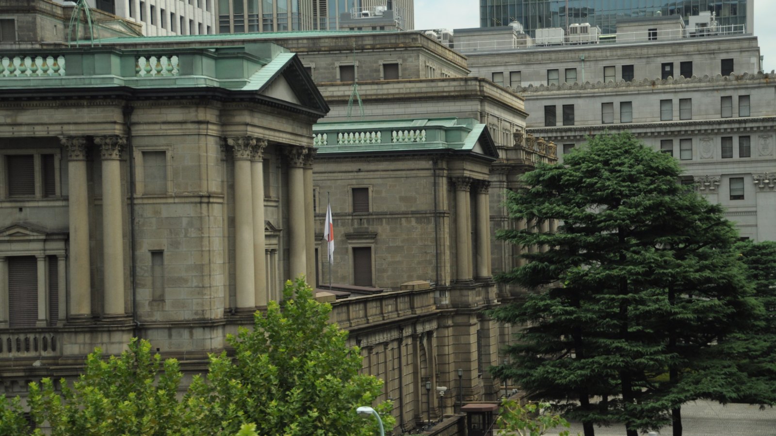 The Bank of Japan head office in Tokyo, with its stone classical buildings, green copper roofs and a Japanese flag among surrounding trees.