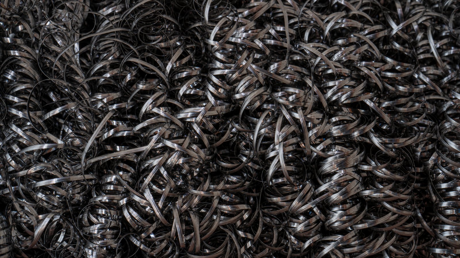 A dense mass of curled steel machining swarf filling the frame, hundreds of bright ribbon-like turnings coiled and overlapping, their edges catching hard light against deep shadow.