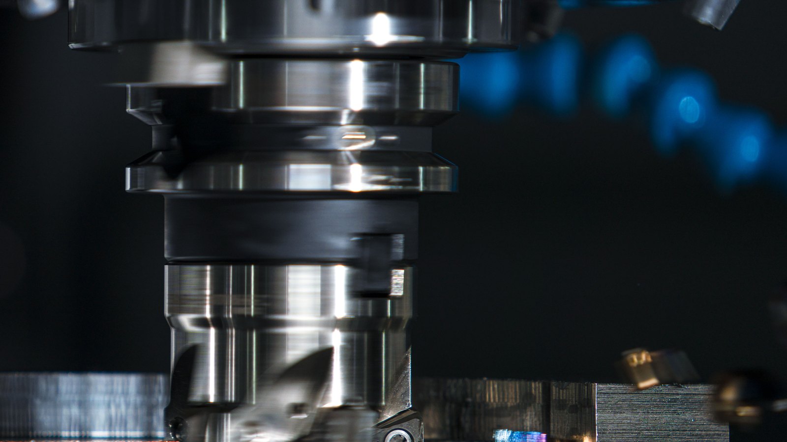 A close macro of a bright steel milling cutter head lowered over a machined metal block, with blue coolant lights blurred in the dark behind it.
