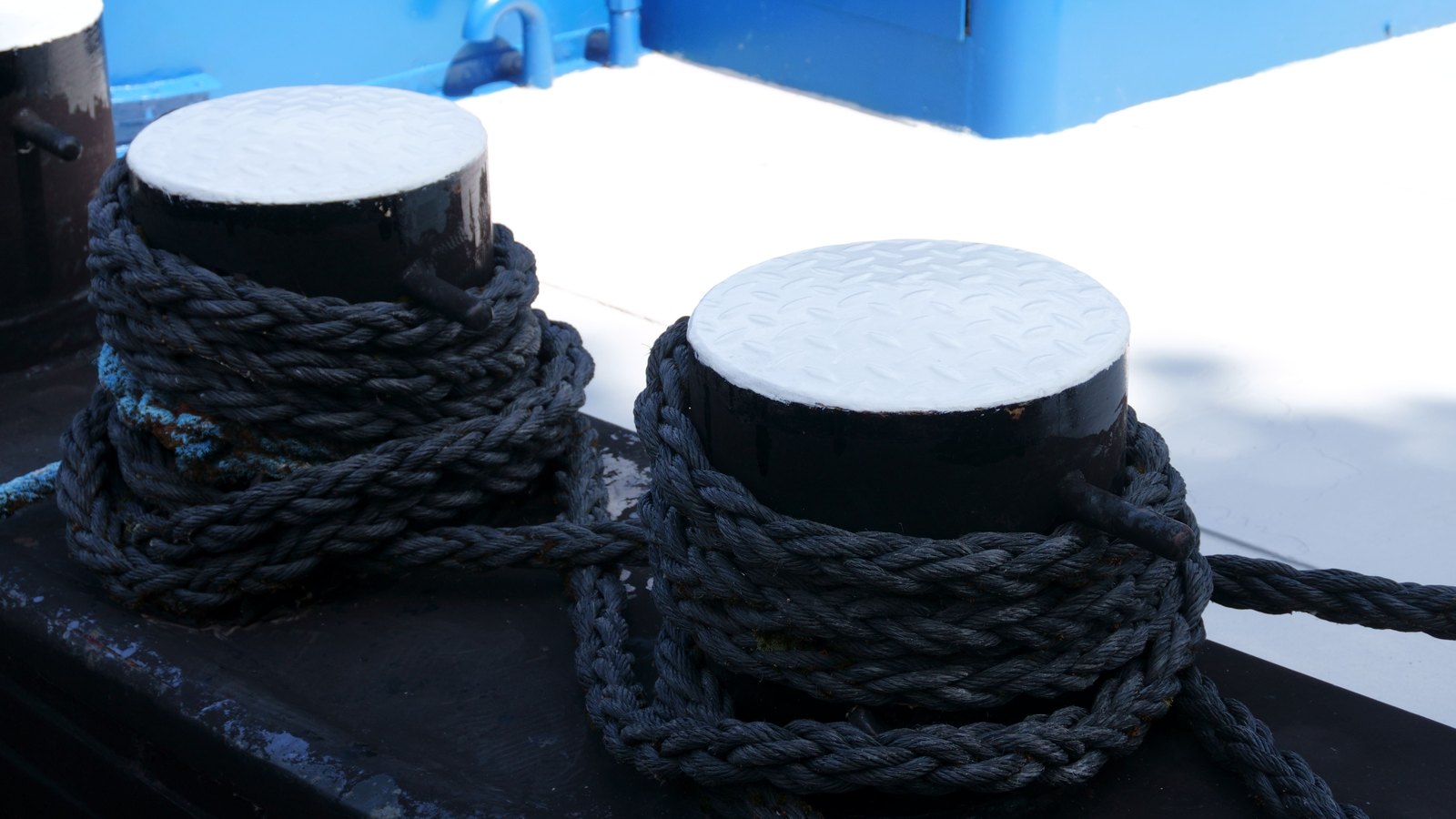 Two black steel mooring bitts with white chequer plate tops stand on the deck of a vessel, heavy grey rope wound in figure of eight turns around both, with a blue painted fitting and white deck behind. An illustrative photograph, not a picture of any cargo counted in these figures.