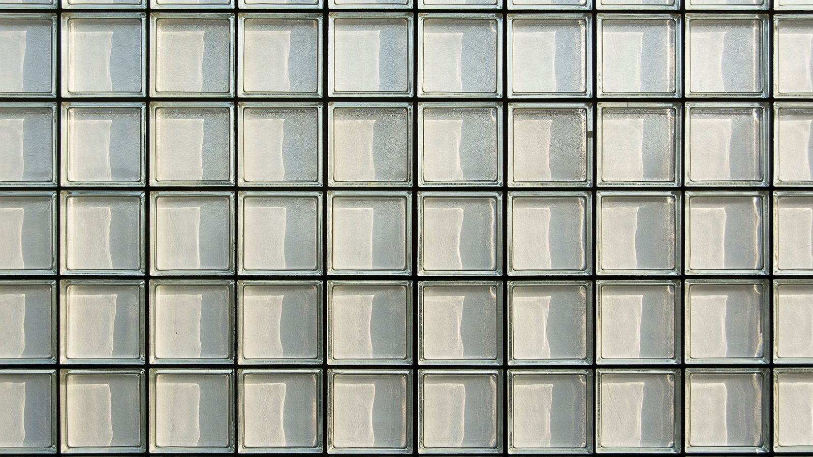 A regular grid of pale square glass blocks filling the whole frame, each block softly reflecting daylight, with thin dark joints between them. No window view, horizon, person, lettering or brand mark is visible.