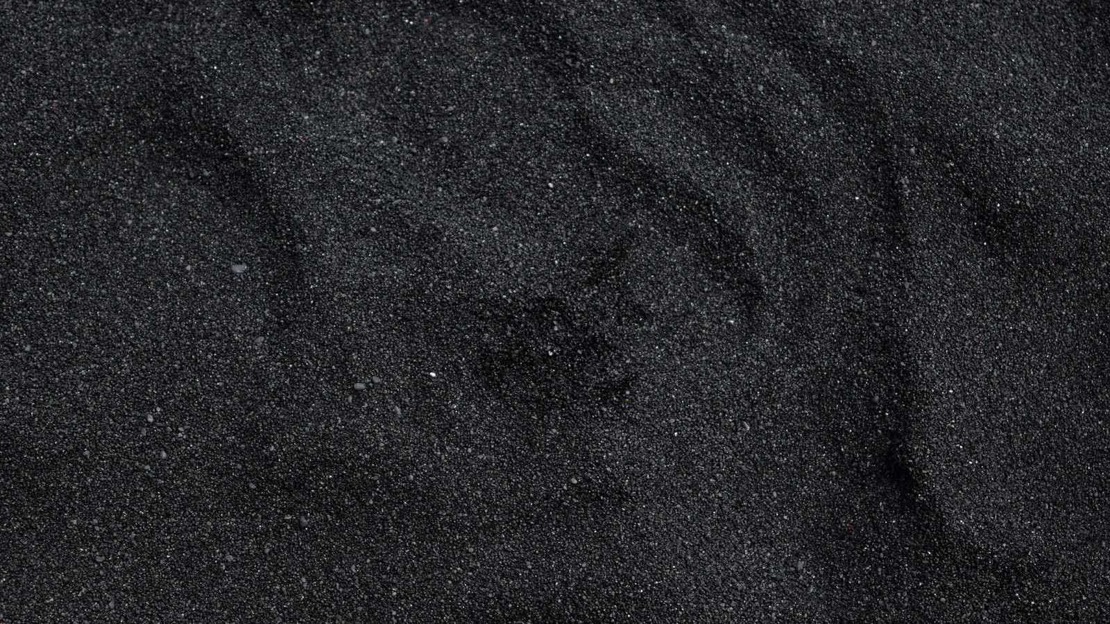 A close photograph of fine black volcanic sand filling the frame, the individual grains sharp and faintly glinting, with soft wind drifts running diagonally across the surface.