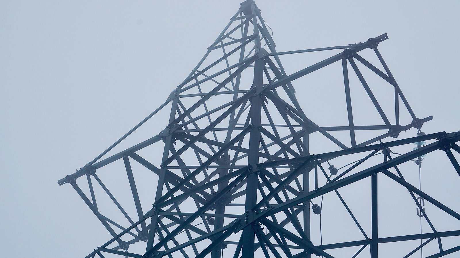 A low angle photograph of the upper part of a steel lattice electricity transmission tower against a flat pale grey sky. Cross braced angle iron members converge at the peak, bolted plates and small insulator fittings hang from the lower arms, and the far side of the structure fades into haze. No text, marking, brand, person or place is in view.