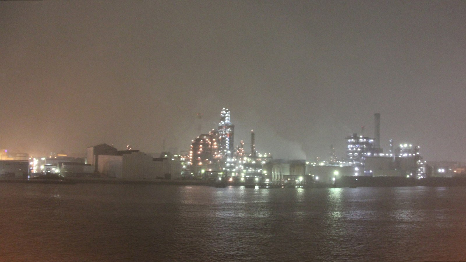 Lit chemical plants and refinery towers of the Keihin industrial zone in Kawasaki seen across the water on a misty evening.