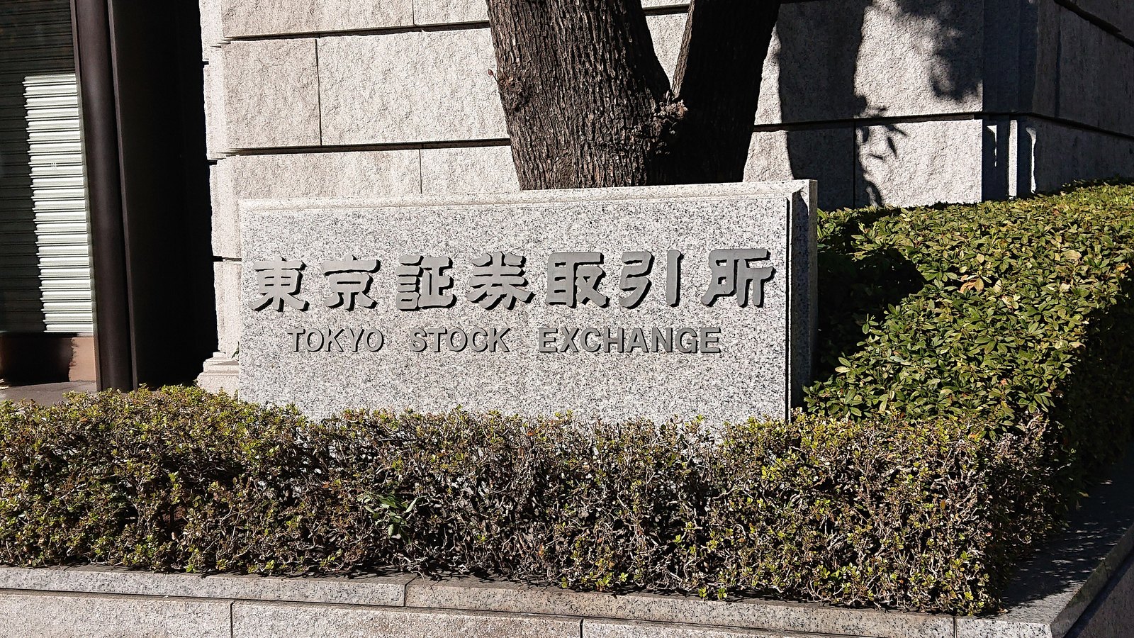 The stone Tokyo Stock Exchange name marker outside the exchange's main building, framed by hedges and a tree.
