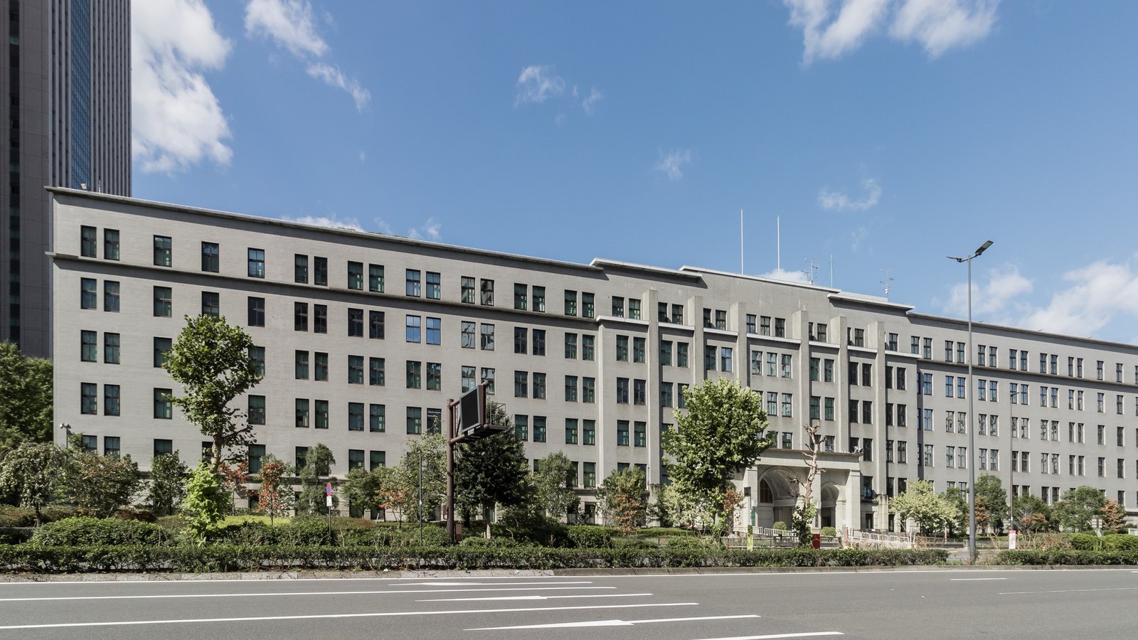 The long grey stone facade of the Ministry of Finance building in Kasumigaseki, Tokyo, behind street trees and an empty multi-lane road under a blue sky.