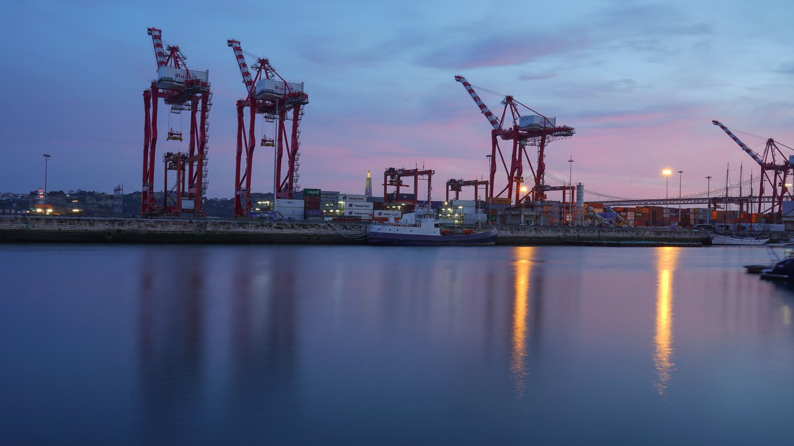 Container cranes stand across calm harbor water at twilight.