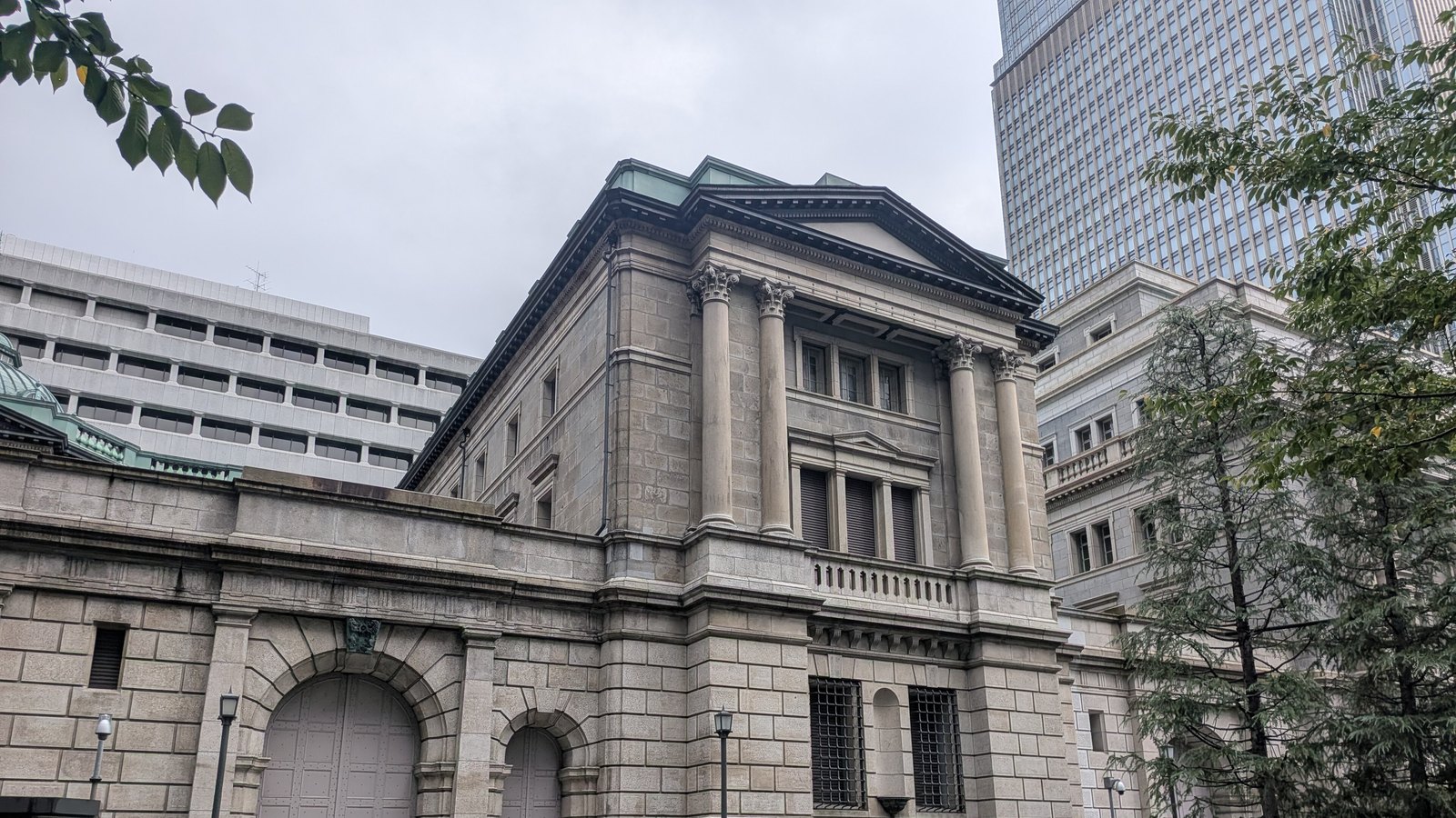 The Bank of Japan head office in Nihonbashi, Tokyo, seen from the street under an overcast sky: a granite neoclassical block with paired Corinthian columns and a pediment, an arched carriage gate in the boundary wall, the green copper dome at the far left and a modern glass tower behind.