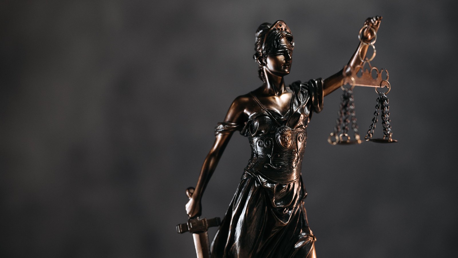 A bronze statuette of Lady Justice, blindfolded, holding a set of scales in one raised hand and a sword in the other, lit against a dark grey background. Stock photo, not a photograph of this event.