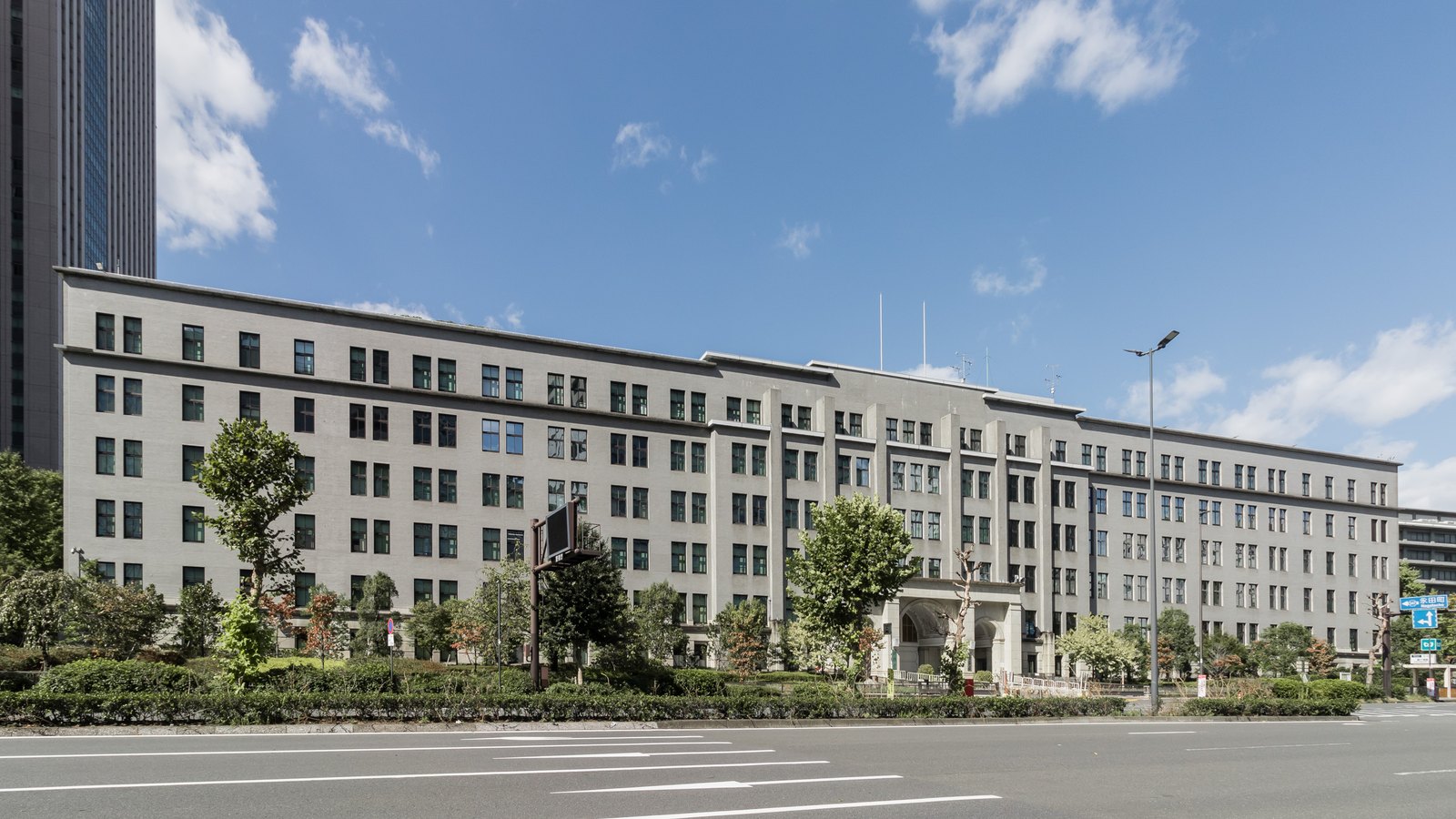 The Ministry of Finance building in Kasumigaseki, Tokyo, photographed square on from across an empty road on a clear day. A long grey stone facade of six storeys runs the width of the frame, with regular rows of small windows, a central arched entrance and low trees planted along the front. A tower block stands at the left edge and the sky above is blue with light cloud.