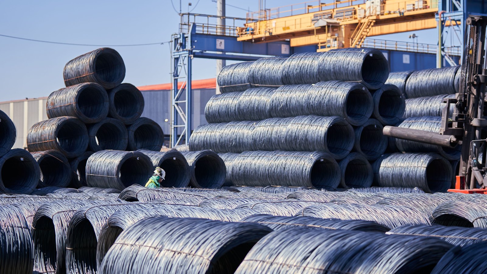 Stacked coils of steel wire rod in an outdoor storage yard, with a gantry crane behind.