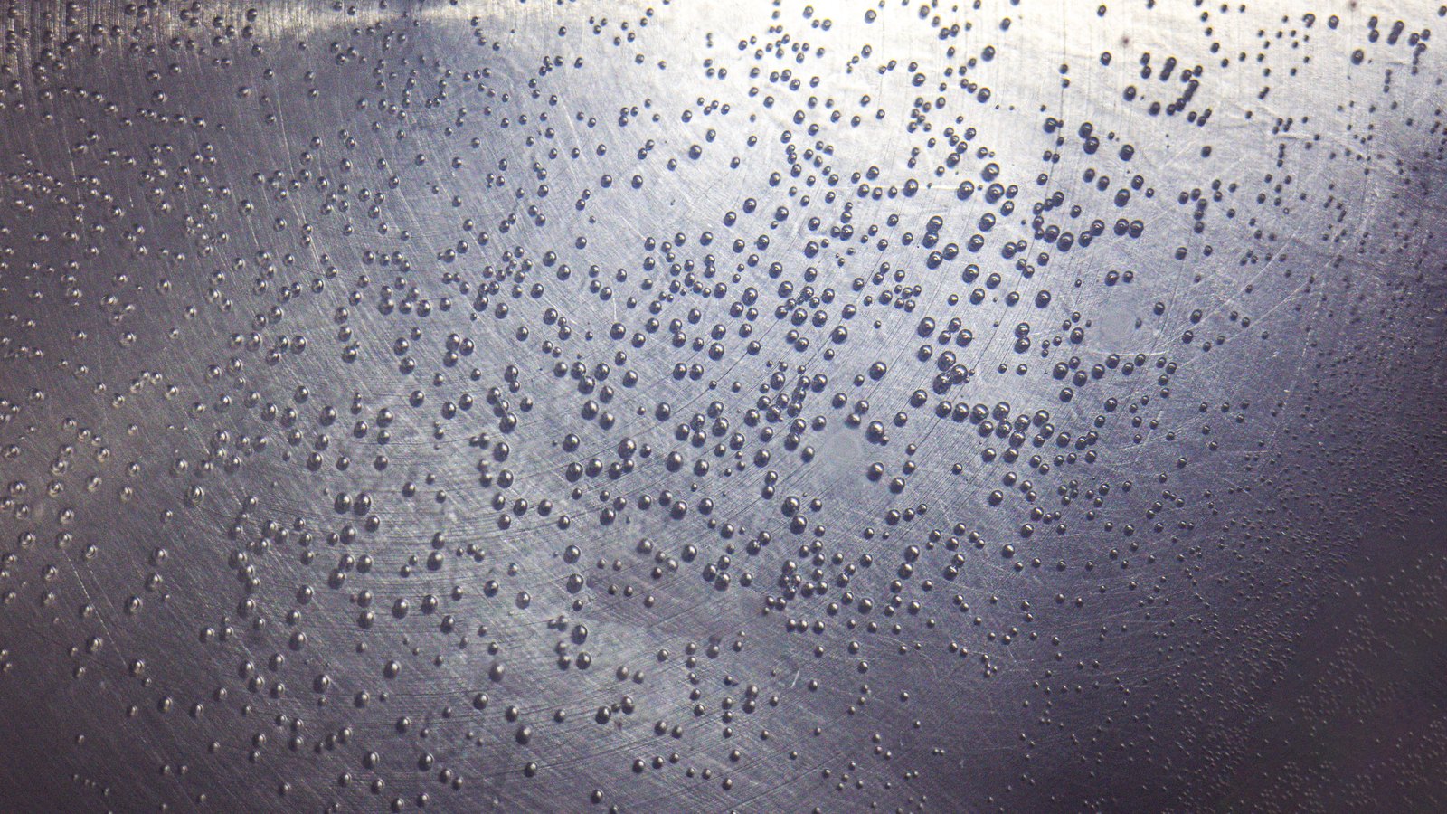 A close photograph of a brushed metal surface scattered with fine beads of condensation, each droplet catching the light against faint circular polishing marks in the metal beneath. The frame is filled edge to edge by the surface and holds no object, person, lettering or brand mark.