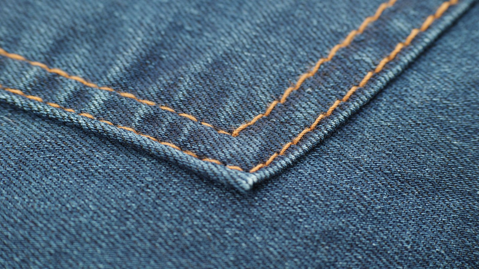 A close photograph of the corner of a denim pocket, the mid-blue twill running diagonally across the frame and two lines of orange topstitching meeting at a point, with no label, rivet or brand mark in view.