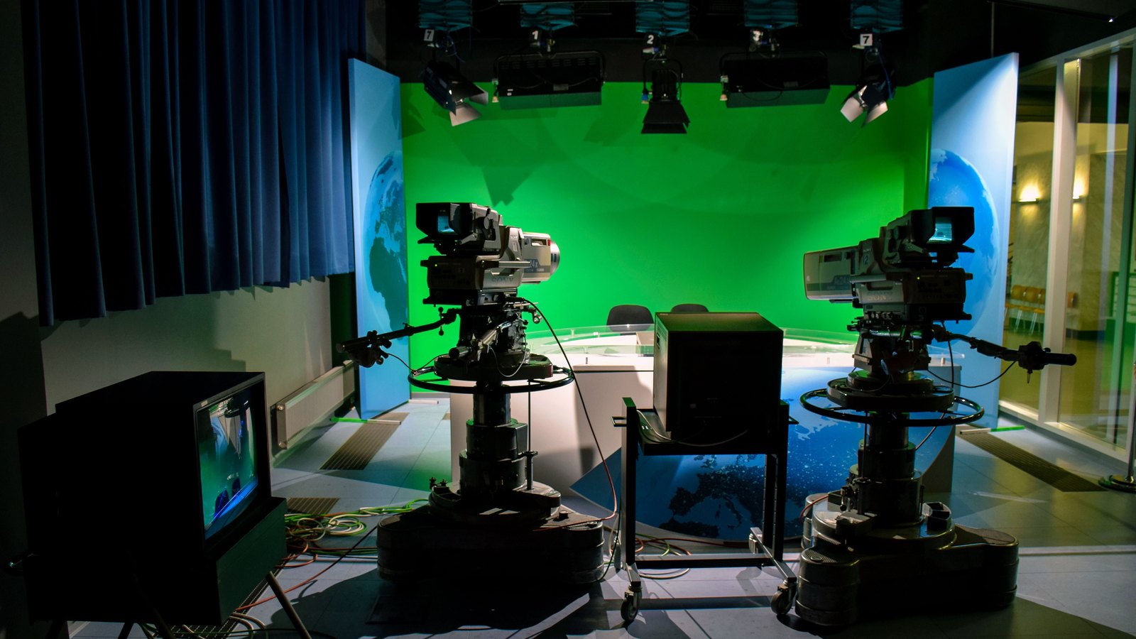 An empty television studio, two pedestal cameras facing a lit green screen and an unoccupied news desk. An illustration, not a photograph of any Telemundo studio, of José Suárez or of the crash.