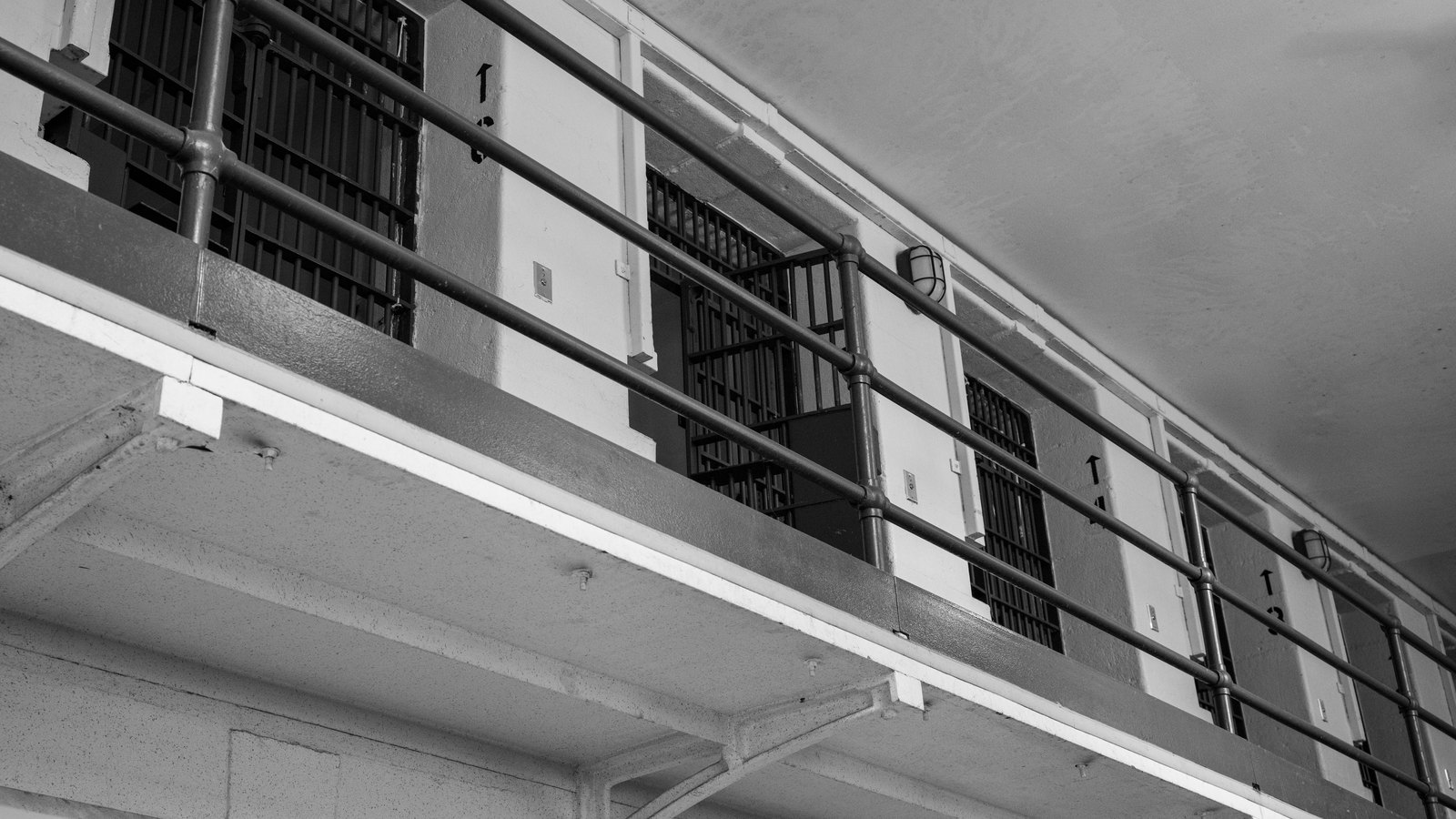 Looking up at an upper tier of a prison cell block in black and white, with a row of barred cell doors set along a walkway behind a pipe railing, no people, signage, text, logos or identifiable location anywhere in the frame. A generic illustration of a prison sentence, not a photograph of this event.