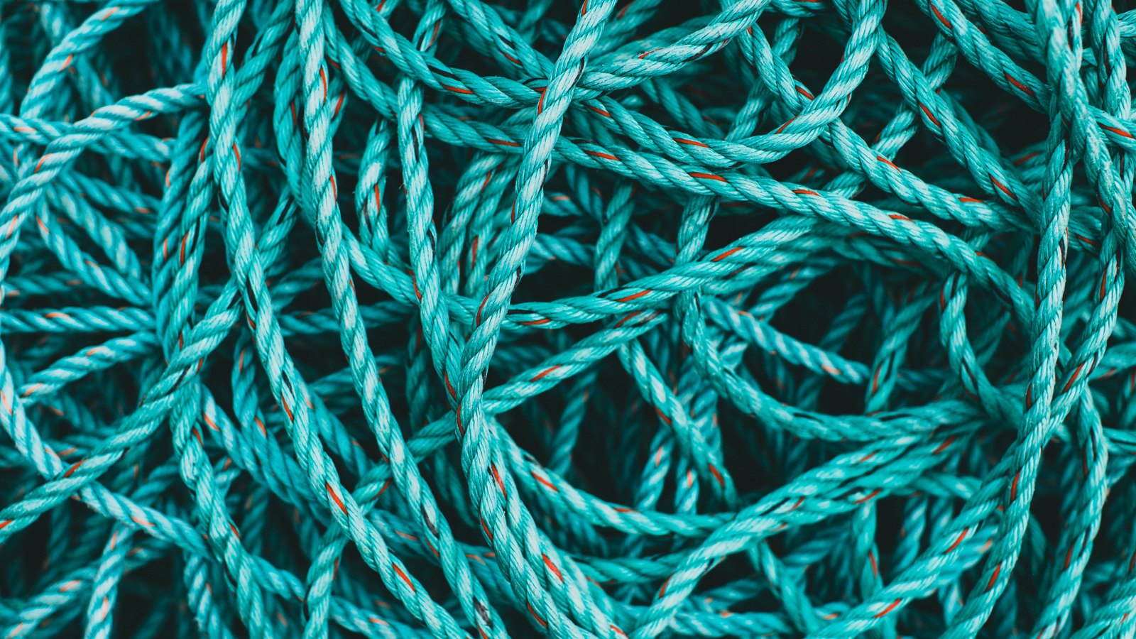 A close photograph of turquoise nylon rope heaped and tangled so that it fills the whole frame, the twist of each strand and a thin orange tracer thread visible along the lengths in front, the rope behind falling into dark shadow. No text, lettering, brand mark, person or place is in view.