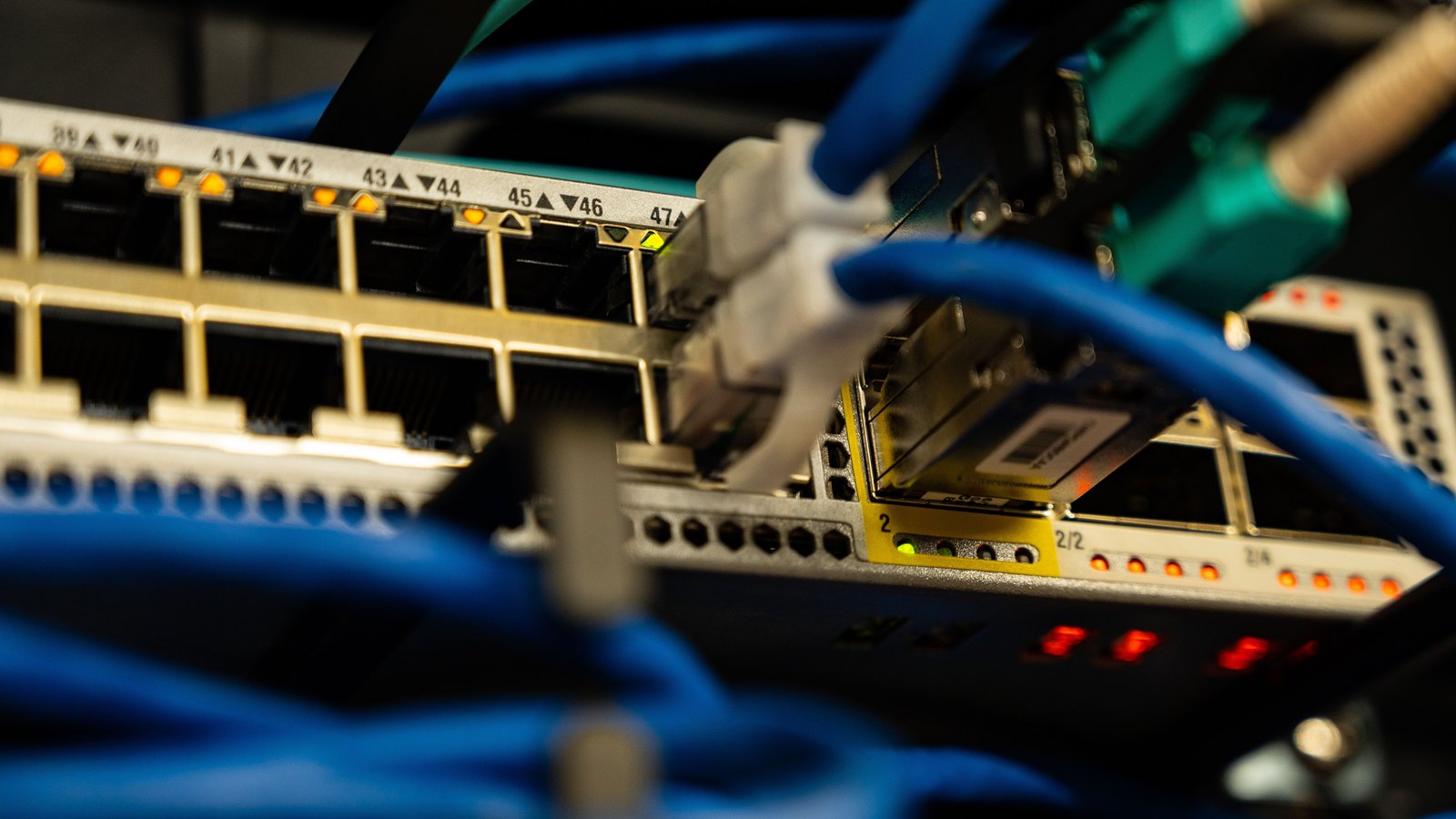 A close view of the front of a network switch. Blue patch leads run into a row of numbered ports, the port legends reading 39 and 40 through to 47, with small amber and green indicator lights above them and a second switch out of focus behind. No brand, logo, company name, person or place is in the frame.