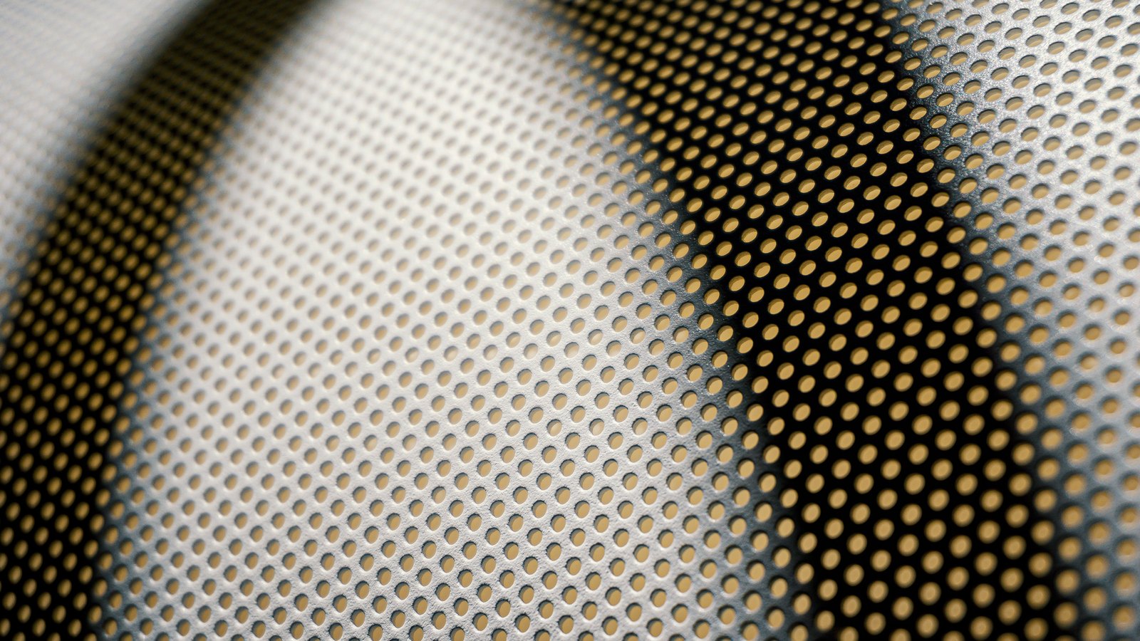 A close crop of a gently curved sheet of perforated metal, its regular grid of small round holes filling the frame, with a broad band of light falling across the middle and darker shadow to either side.