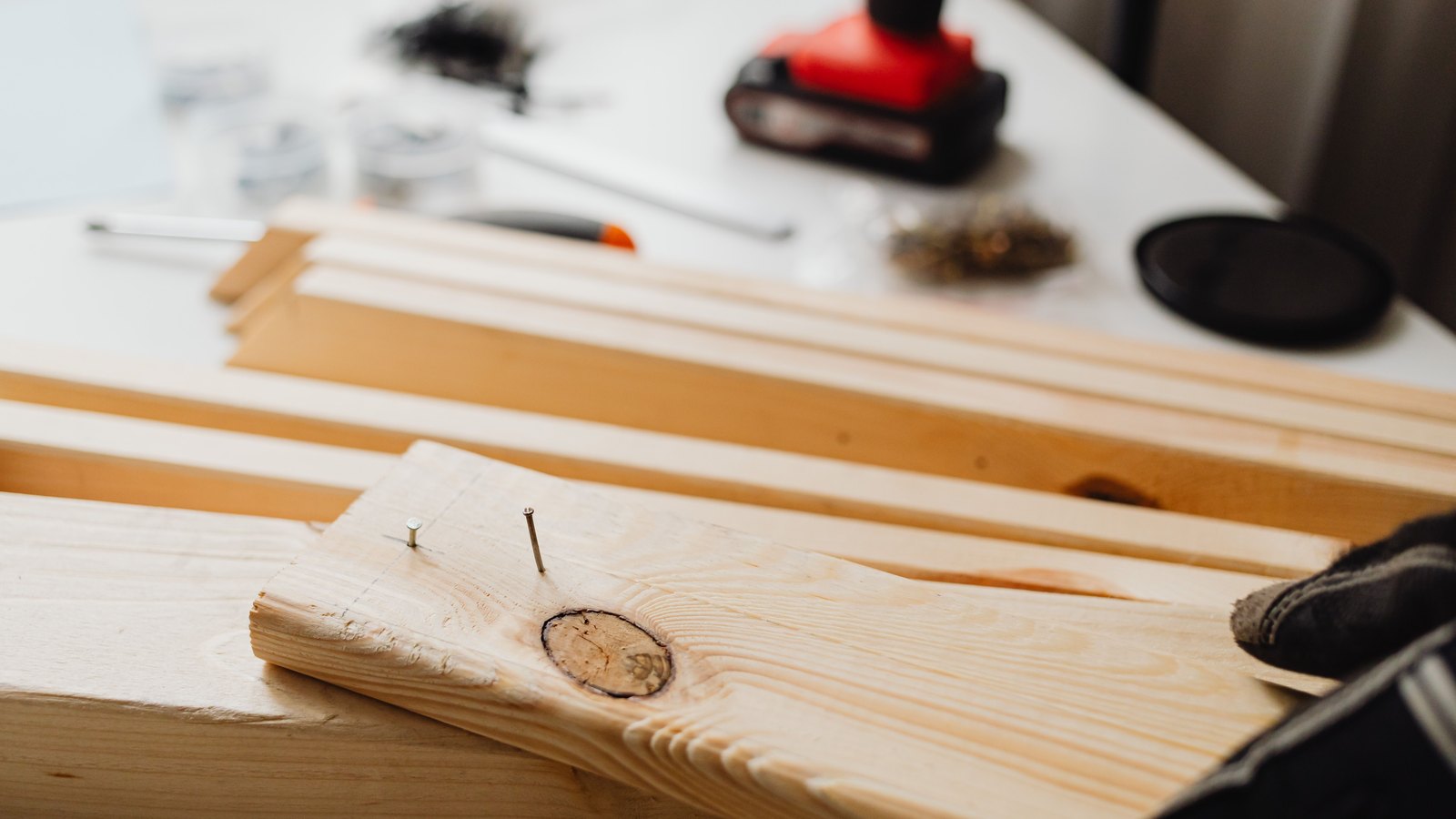 A close view of pale untreated pine boards laid across a workbench, with two thin nails part driven into the nearest board beside a knot in the grain. Behind them, out of focus, are a white work surface holding a pencil, a utility knife, small jars of fixings and a red cordless tool battery, with a gloved hand entering the frame at the right edge. No faces, buildings, signage or legible lettering appear. A generic illustration of construction work, not a photograph of any Kelly Roofing job or of any customer's property.