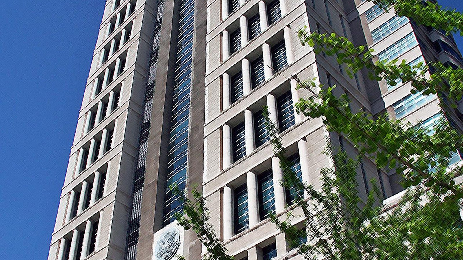 The Thomas F. Eagleton United States Courthouse in St. Louis seen from street level looking up, its pale limestone piers and recessed windows running to the top of the frame against a hard blue sky, a carved circular medallion low on the wall and a leafy tree filling the right of the picture.