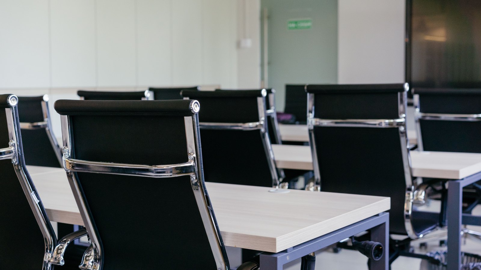Rows of empty black leather and chrome chairs pushed up to pale wooden desks in a bright, unoccupied meeting room. Nobody is in the frame, and the only sign on the far wall is a small green exit marker too blurred to read. It is a stock photograph of an empty meeting room and not a photograph of any Florida state office.