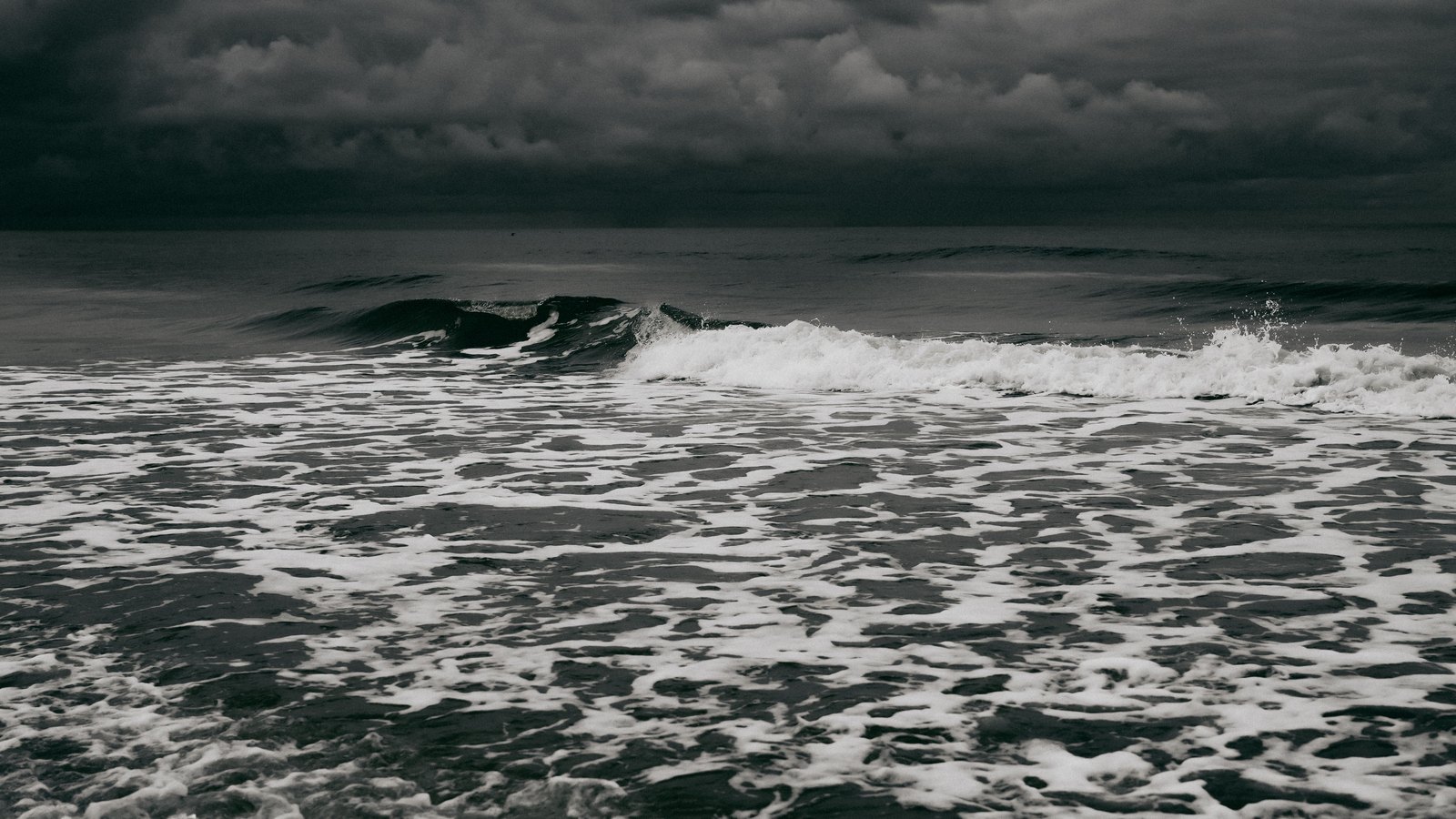 Grey open water under a heavy overcast, a single swell rolling in across a field of white foam, with no land or vessel in sight.