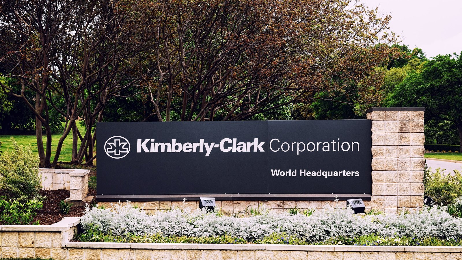A black Kimberly-Clark Corporation World Headquarters monument sign set in a stone planter with silver foliage, trees behind, in Irving, Texas.