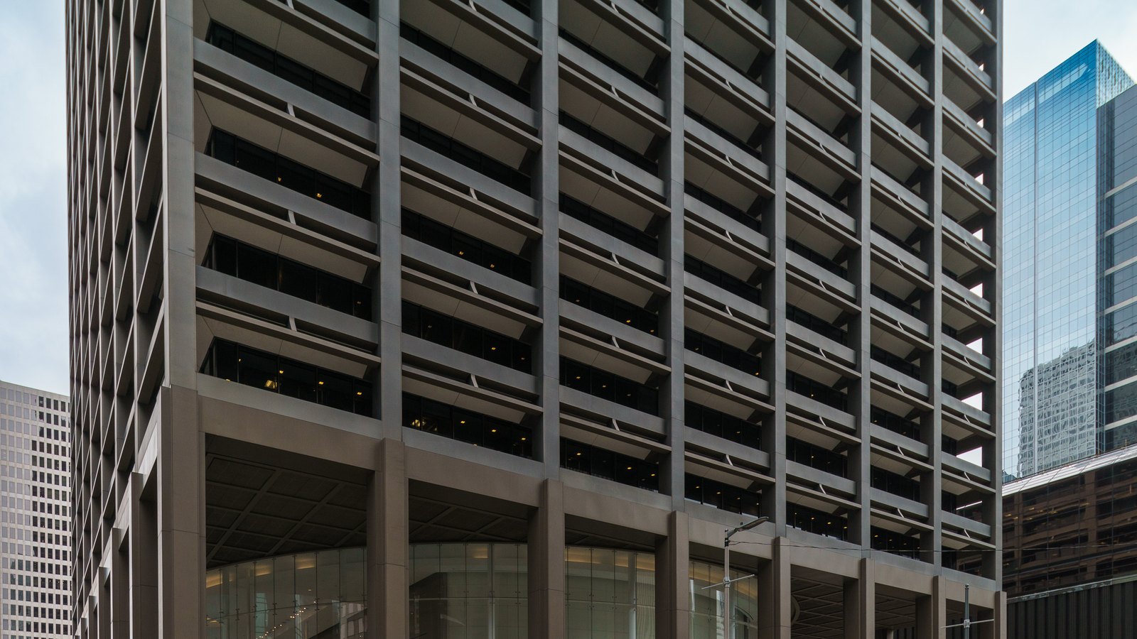 The dark bronze grid facade of the Kinder Morgan Building in downtown Houston, its deep window bays rising above a glass lobby set behind tall columns, with a blue glass tower to the right.