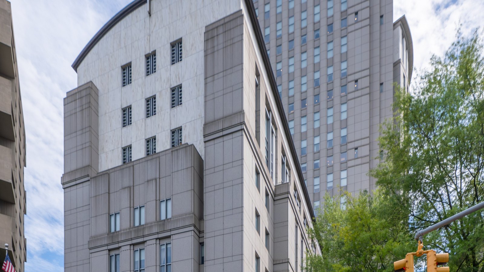 A grey stone federal courthouse in Manhattan photographed from street level looking upward: a curved-topped block faced in pale marble stands in front of a taller tower of small square windows, a leafy tree and a traffic signal fill the right edge, and a United States flag hangs from a neighbouring building at the lower left. No signage or lettering is legible anywhere in the frame.