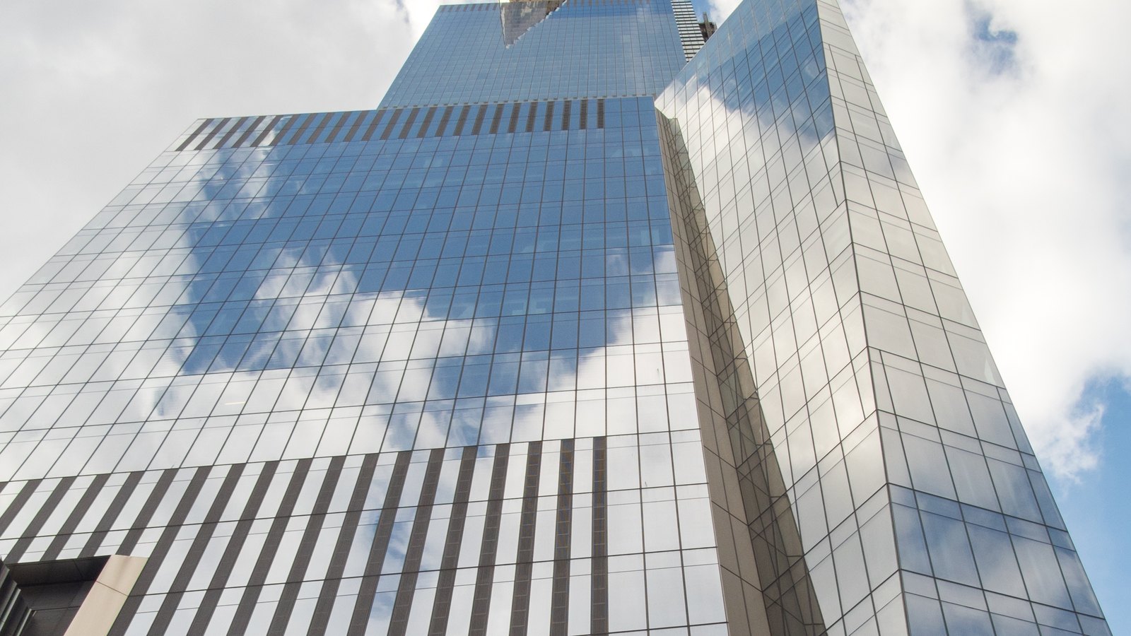 The upper part of 30 Hudson Yards in Manhattan photographed from street level looking straight up, a mirrored glass tower whose facade reflects white cloud and blue sky, with a second glazed elevation running up the right of the frame. No signage or lettering is visible anywhere.