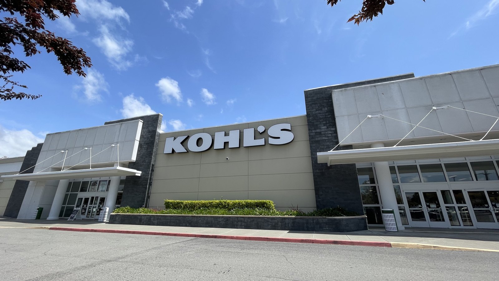 The exterior of a Kohl's store seen from the car park: a low beige and grey building with the KOHL'S wordmark in large white letters across the centre of the facade, glazed entrances at either end, a clipped hedge along the front and an empty parking bay in the foreground, under a blue sky.