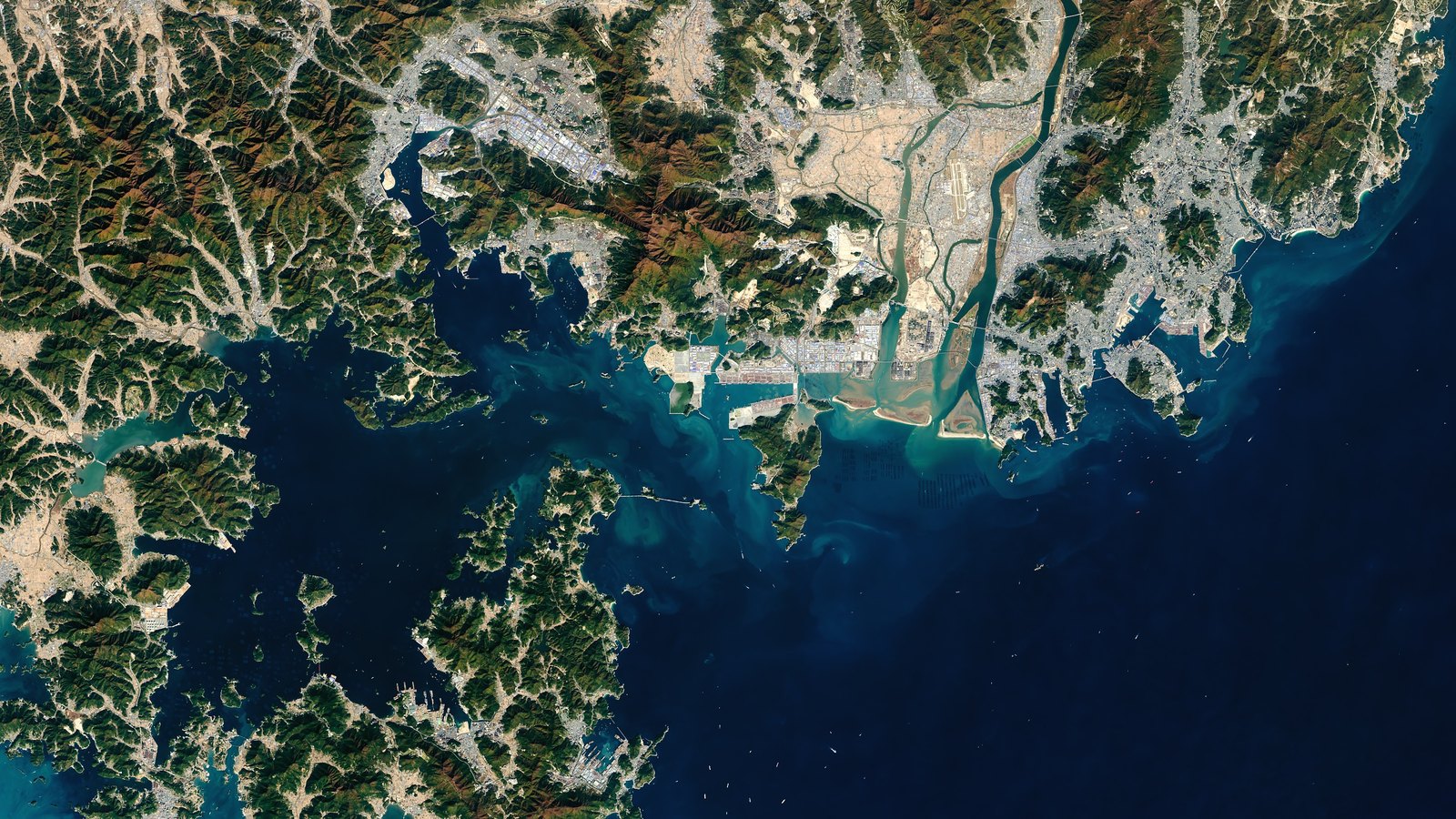 A satellite view of the Port of Busan and the Nakdong River mouth on South Korea's southeast coast, with container terminals on reclaimed land, an airport beside the river and forested hills around a dark blue bay.