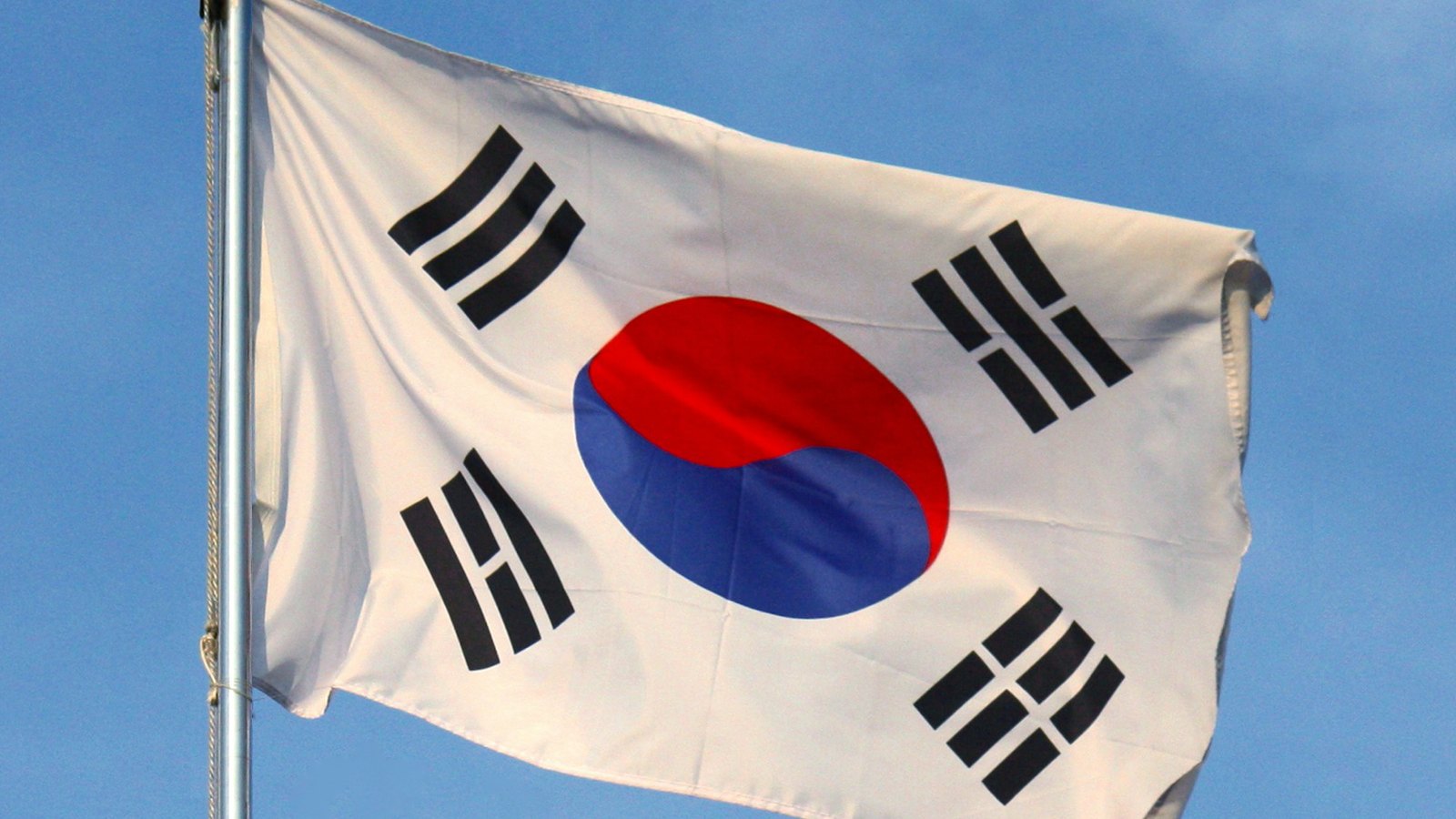 The national flag of South Korea filling the frame, flying from a metal pole at the left edge against a plain blue sky. The white field carries the red and blue taegeuk circle at its centre and a group of three or four black bars at each corner, and the fabric is lifted and creased by the wind. No text, buildings or people are in the frame.
