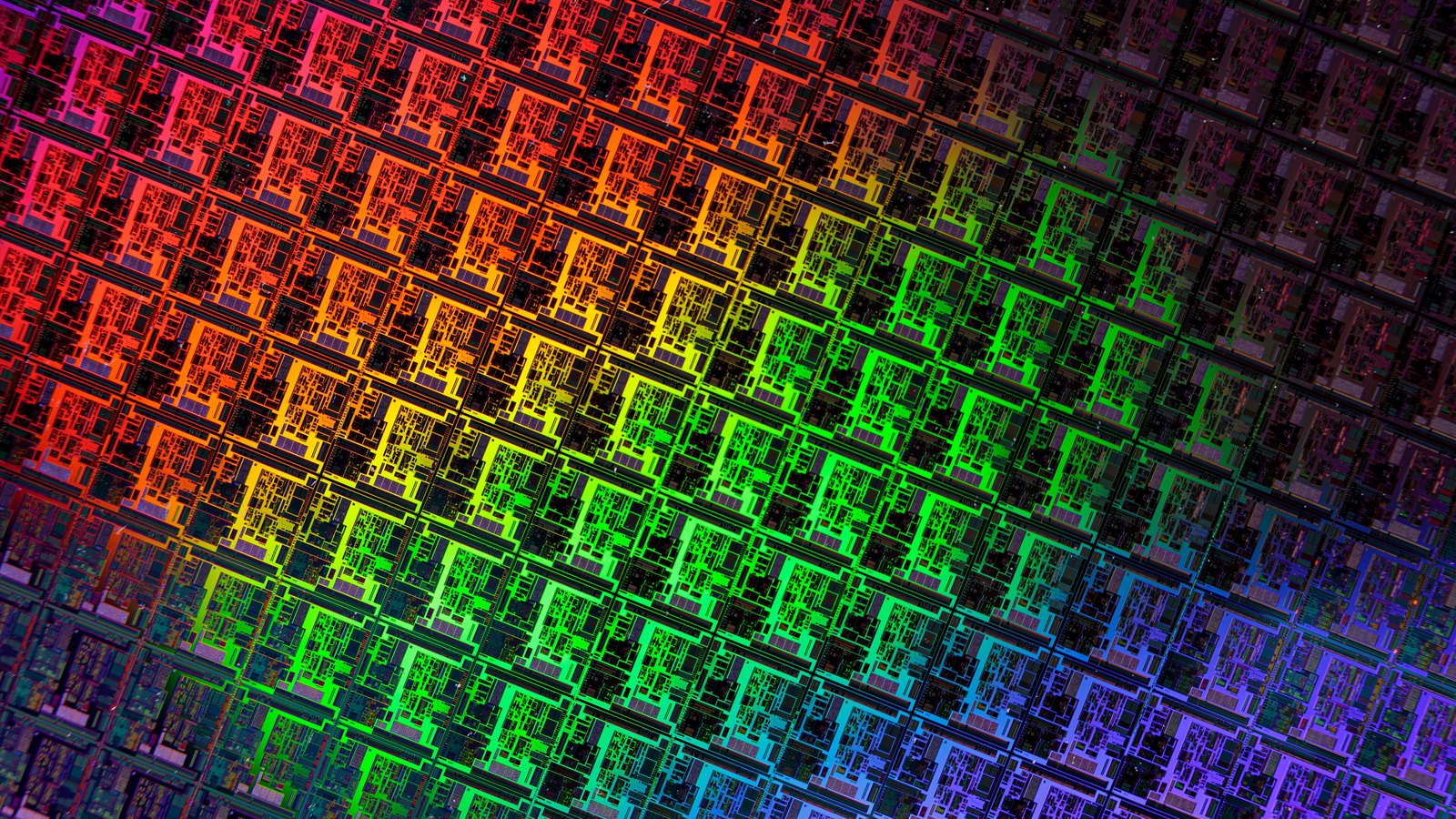 A close crop of a finished silicon wafer, filling the frame with row after row of identical rectangular integrated circuits. Diffraction from the surface throws the grid into bands of magenta, orange, green and blue running diagonally across it. No text, packaging, hand or tool is in the picture.