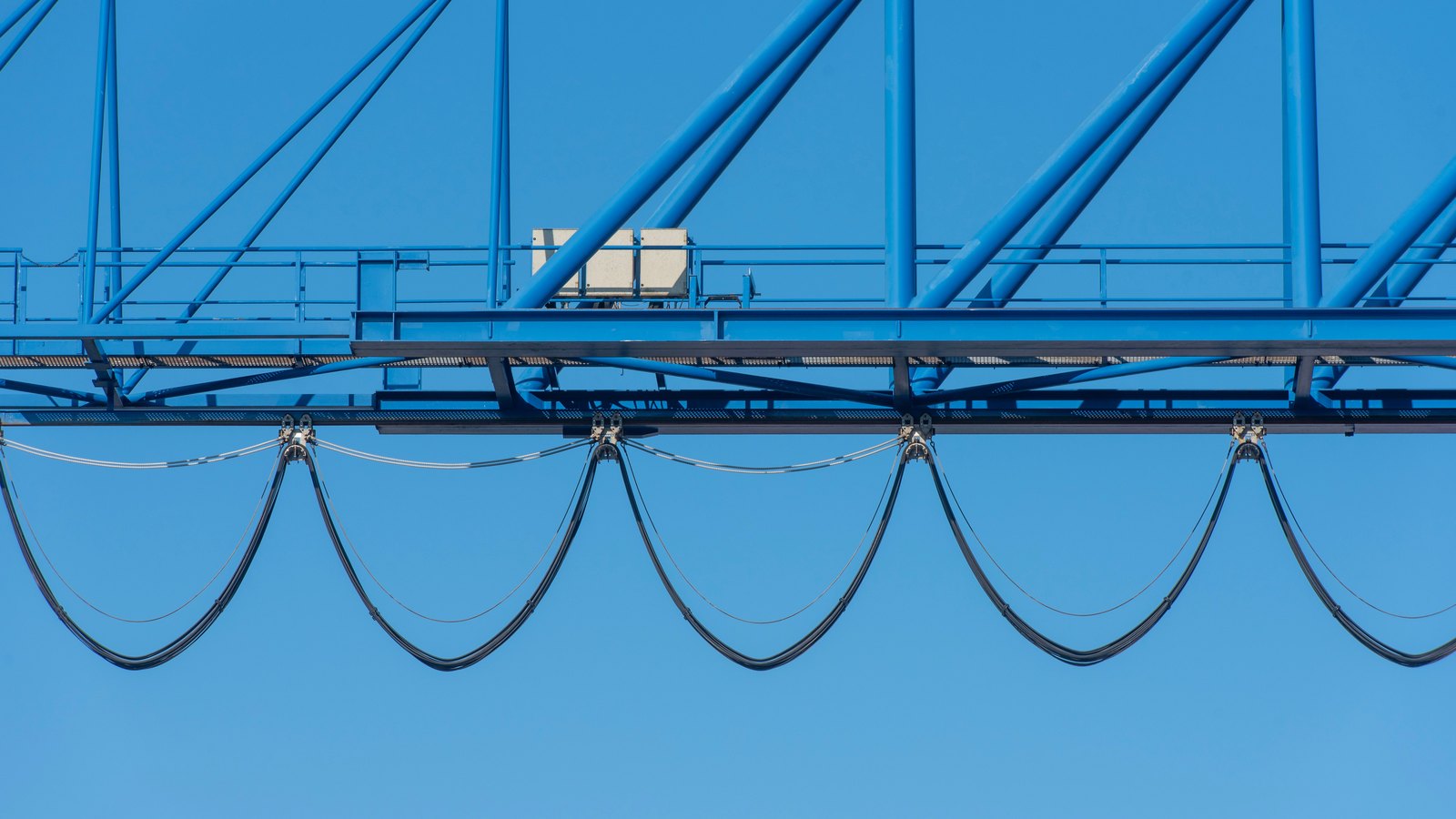 A blue industrial crane beam crosses a clear sky above five deep loops of suspended black cable.