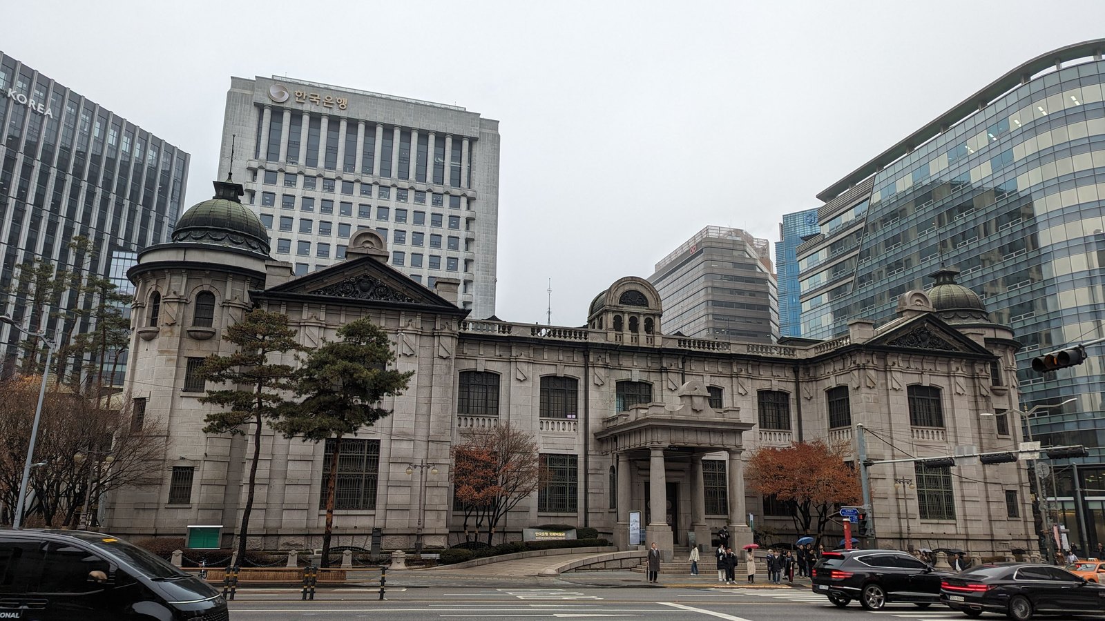 The Bank of Korea Money Museum, the central bank's granite former head office in Seoul, under an overcast sky, with the bank's modern head office tower carrying its name rising directly behind it.