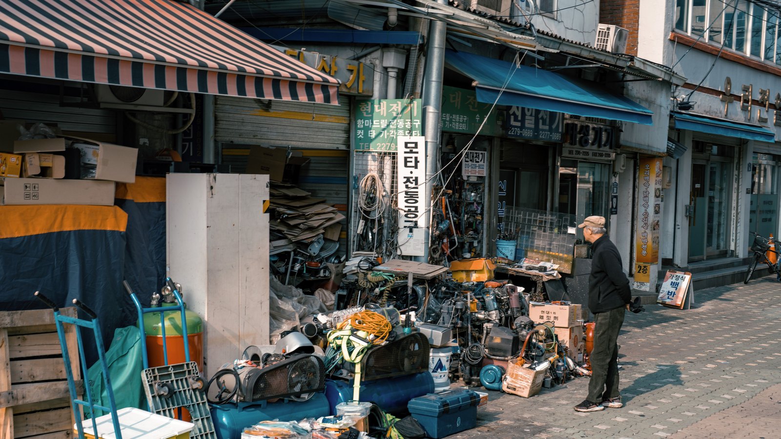 A man stands beside stacks of machinery and tools outside storefronts on a Seoul shopping street.