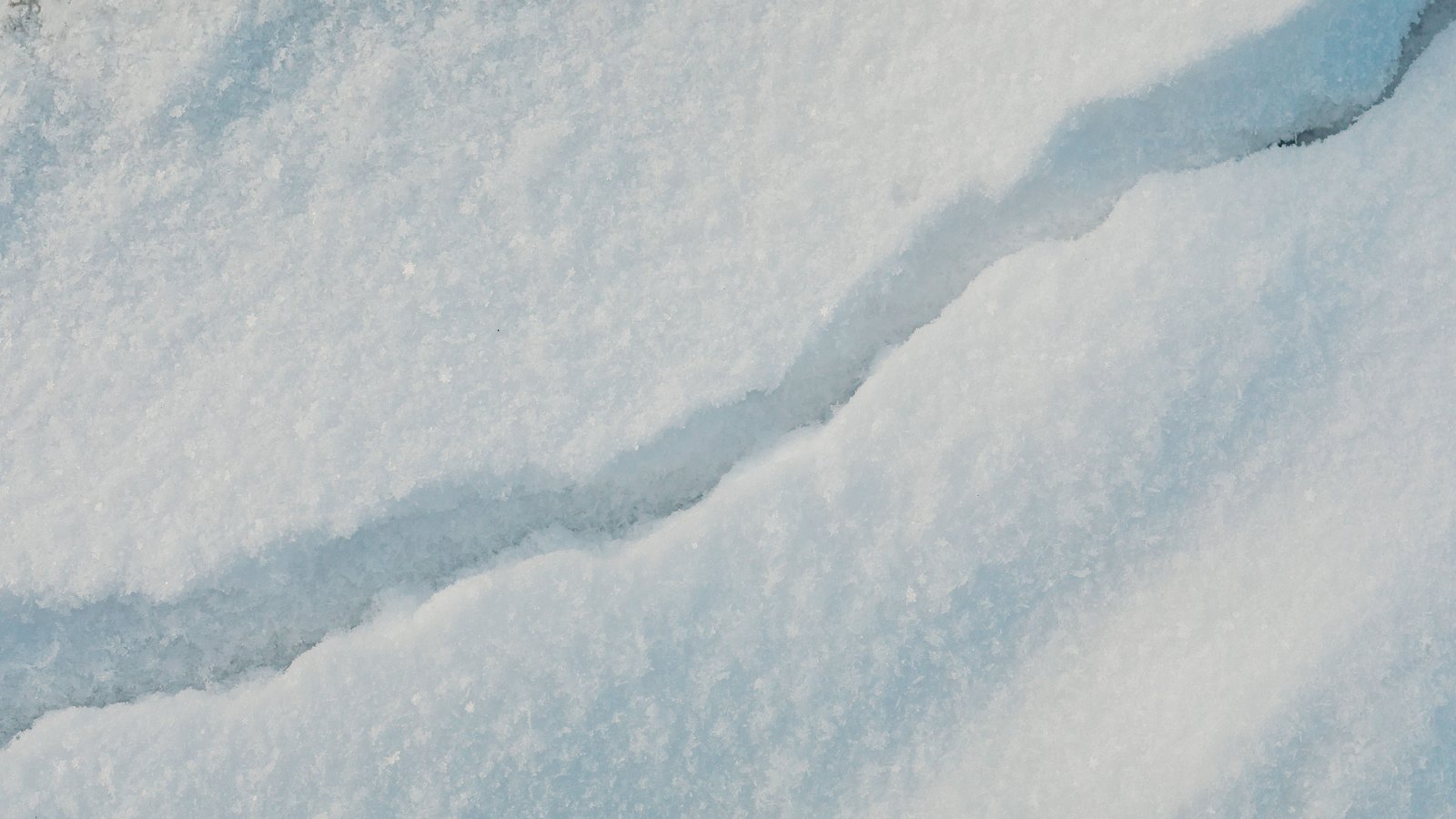 A close macro of a snow surface in pale blue light, split from lower left to upper right by a single deep fracture, with fine ice crystals catching the light along both edges of the break.