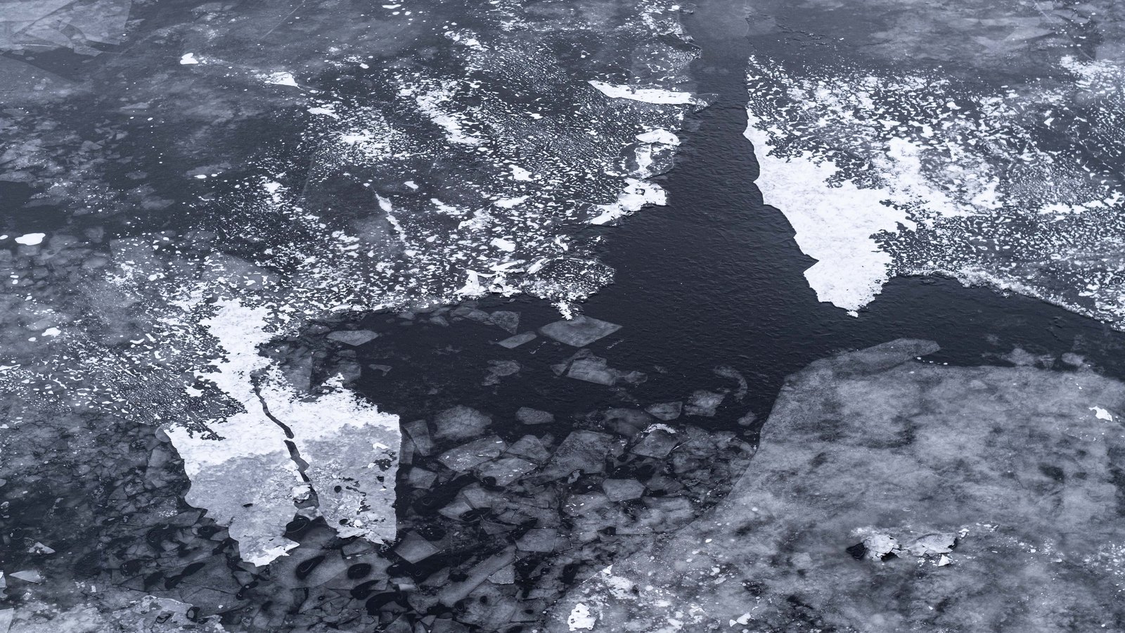 An aerial view of broken sea ice, pale floes and slush separated by a channel of dark open water running through the middle of the frame.