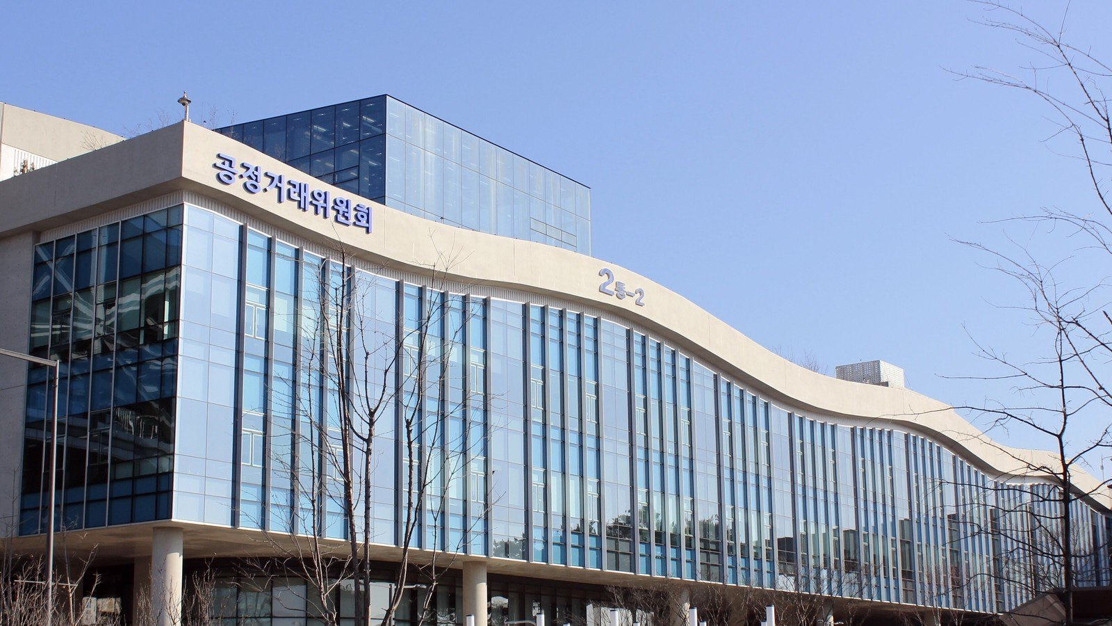 The Korea Fair Trade Commission's building at the Sejong government complex, a curving glass and pale stone facade carrying the commission's Korean name and the building number, behind bare winter trees under a clear blue sky.